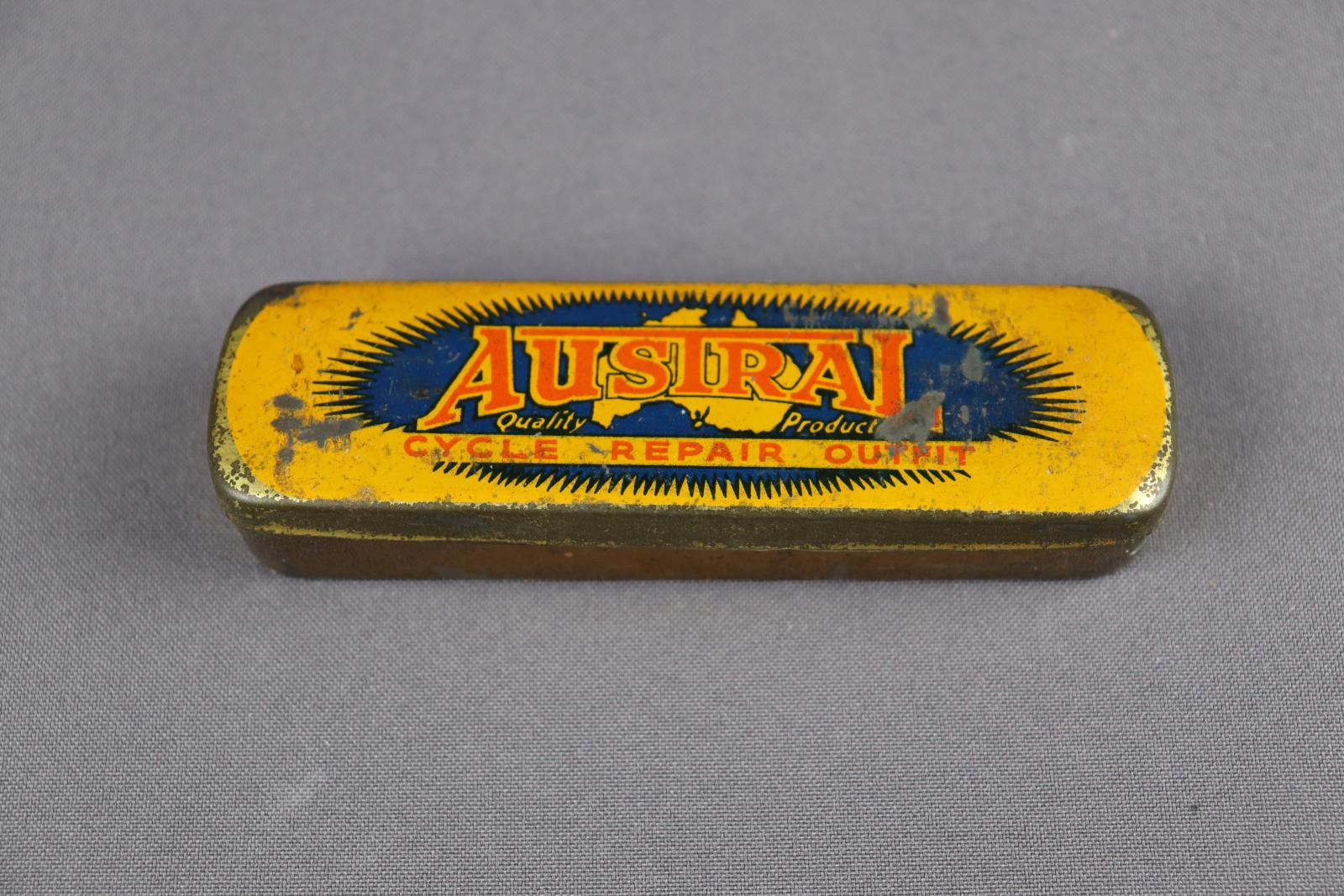 Rectangle metal tin. Lid of tin is yellow with orange text saying Austral Cycle Repair Outfit