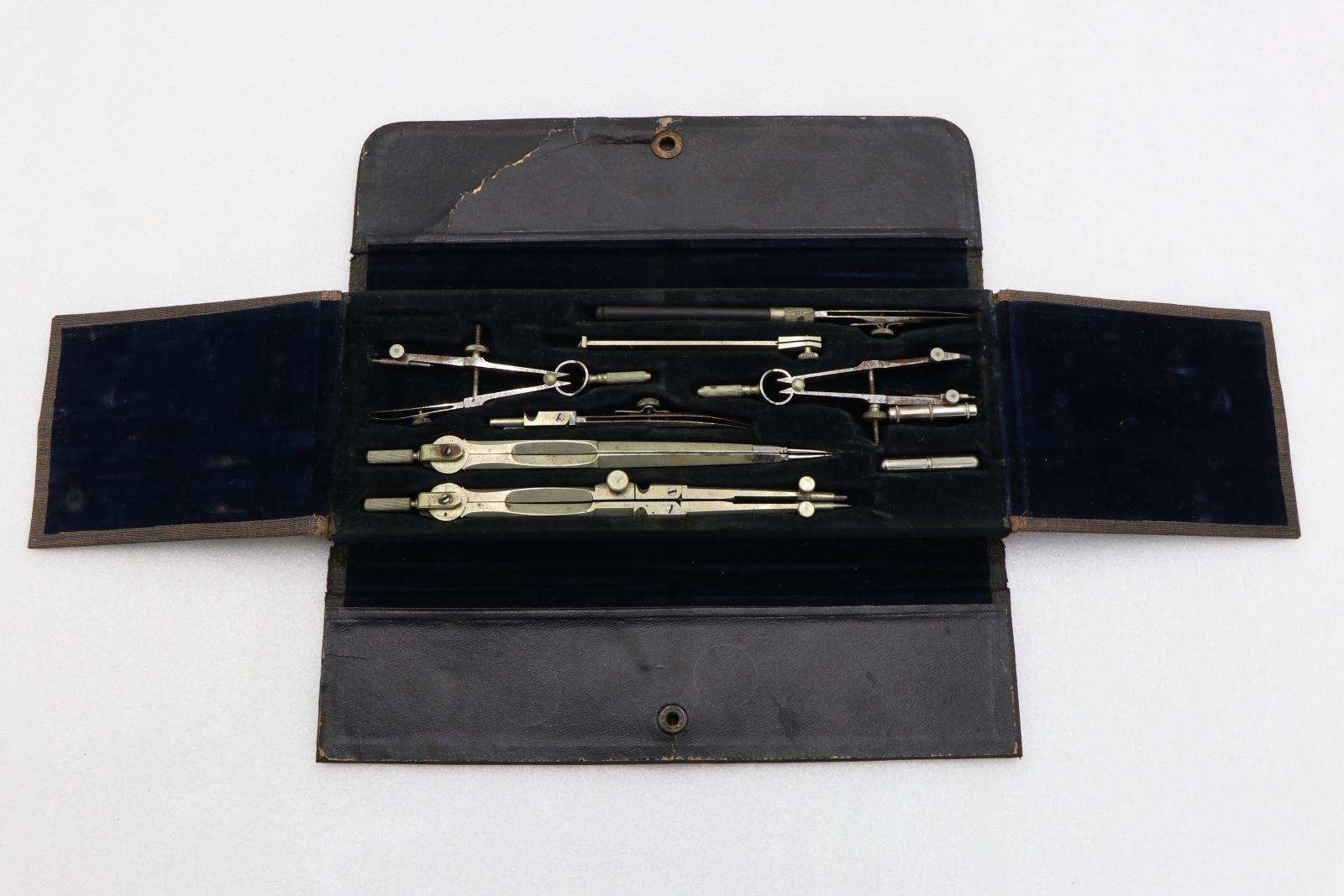 Black rectangular case displayed open, showing seven silver metal instruments for drawing maps and two silver metal tubes for spare lead. Instruments are set in recesses in blue velvet that lines case.