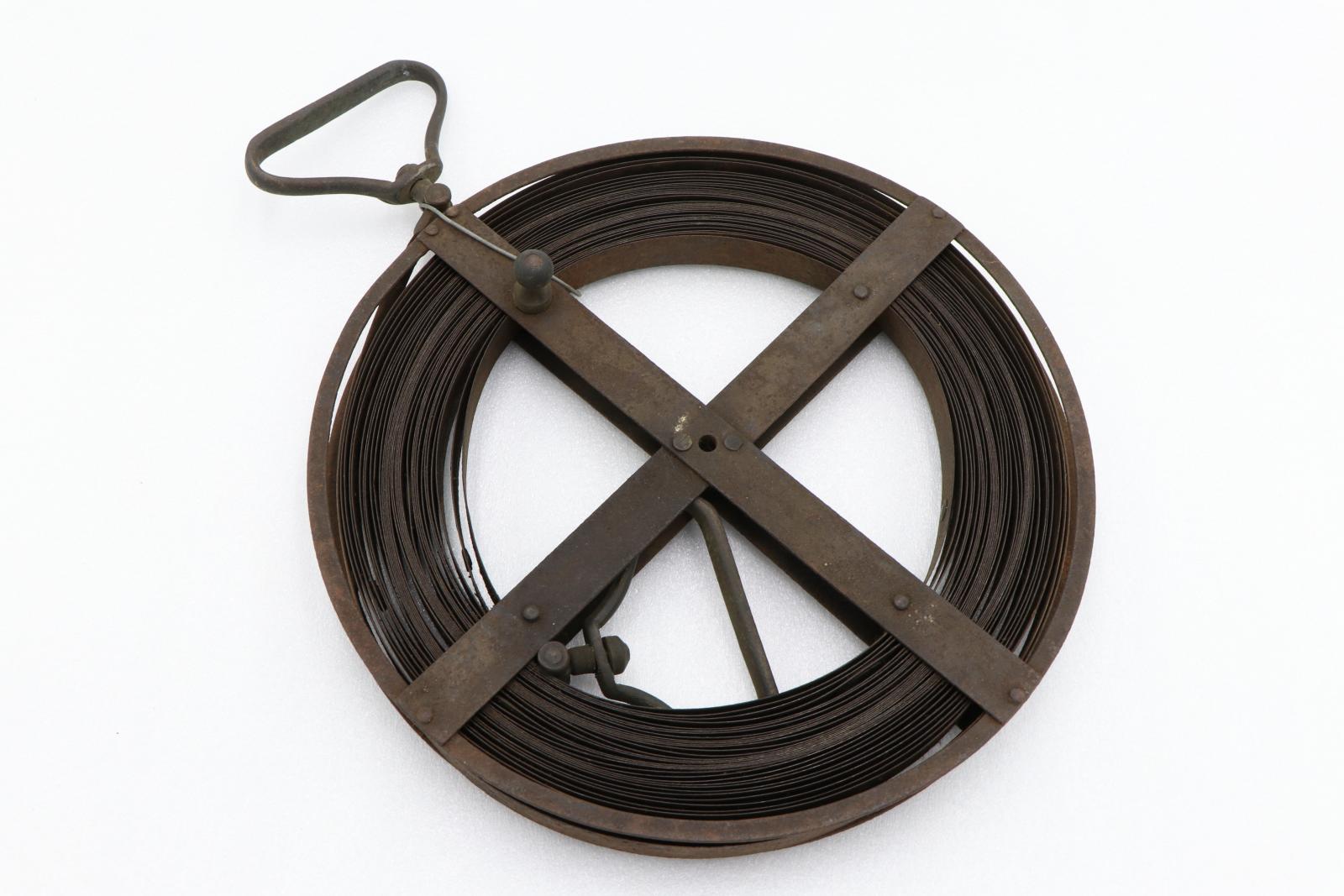 Thin steel tape rolled up on dark metal wheel reinforced with bars to create an X across. Trianglar metal handle on one end of tape visible within wheel. Triangular handle on other end of tape protrudes from top of wheel.
