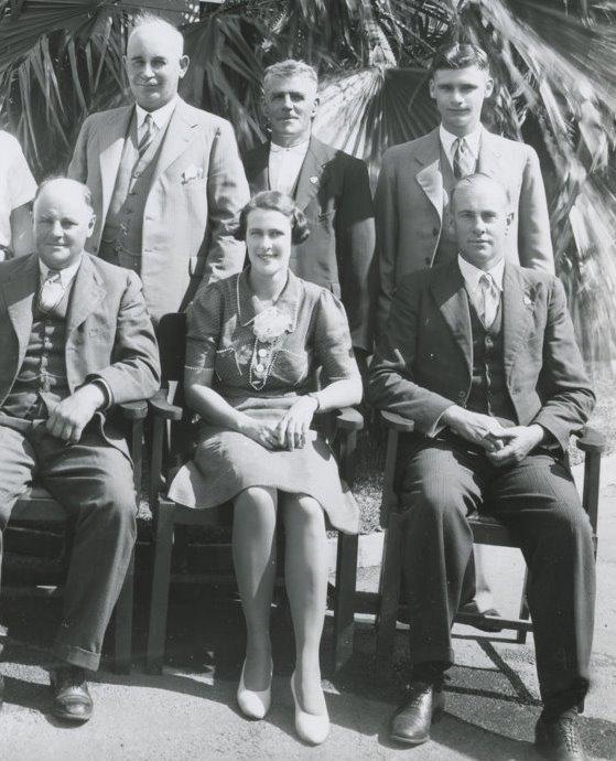 Two rows of people, three standing behind three sitting. In the middle of the front row is a young woman in a dress with short hair, the rest of the people or older men wearing suits. 