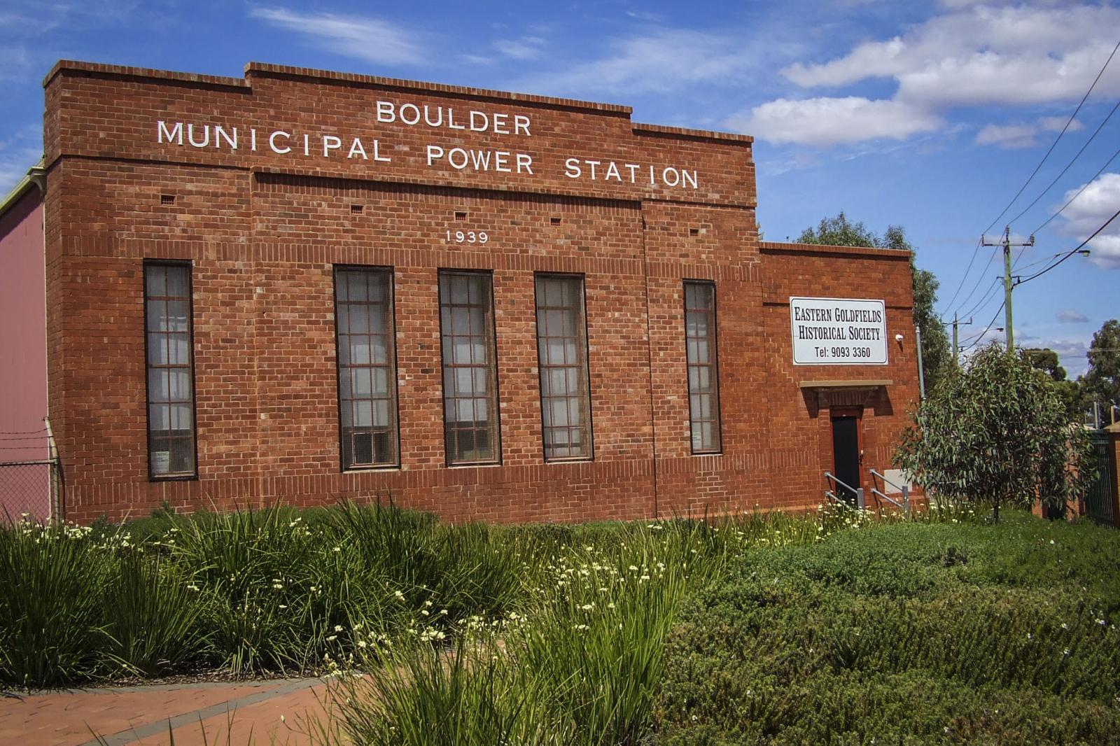 A rectangular red brick building with white lettering "BOULDER MUNICIPAL POWER STATION" across the top. To the right, above the door, is a sign reading "Eastern Goldfields Historical Society". In the foreground are green gardens either side of a brick path.