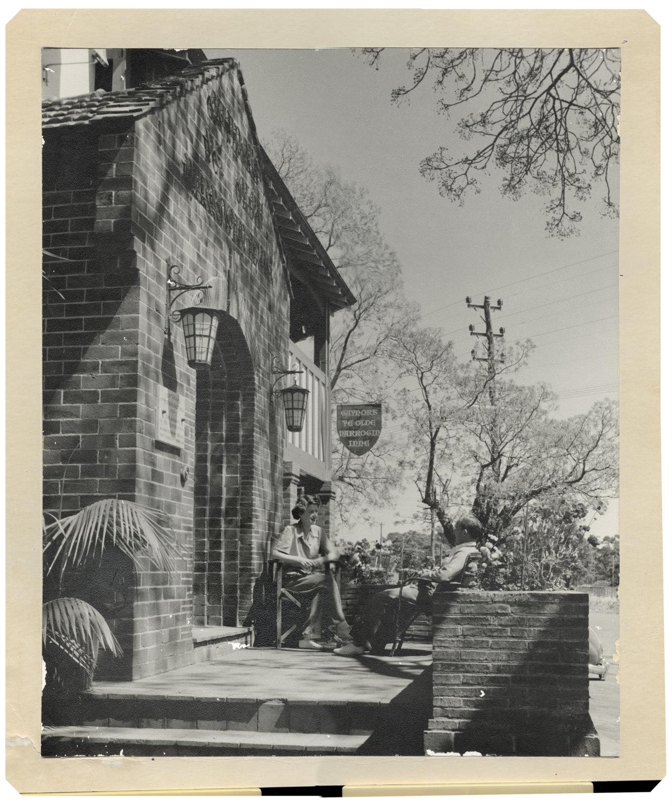 A man and a women sitting outside on a brick patio. To the left is the brick entrance with a metal sigh handing above it.