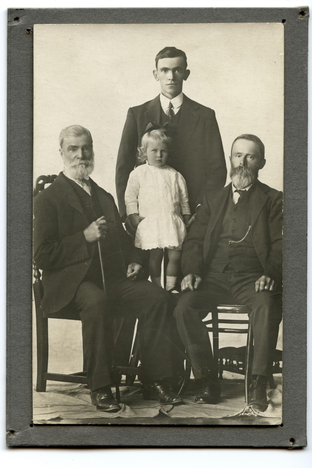 Three men, two sitting, middle man standing all wearing dark suits. In the middle standing on a stand is a female toddler in a white dress.