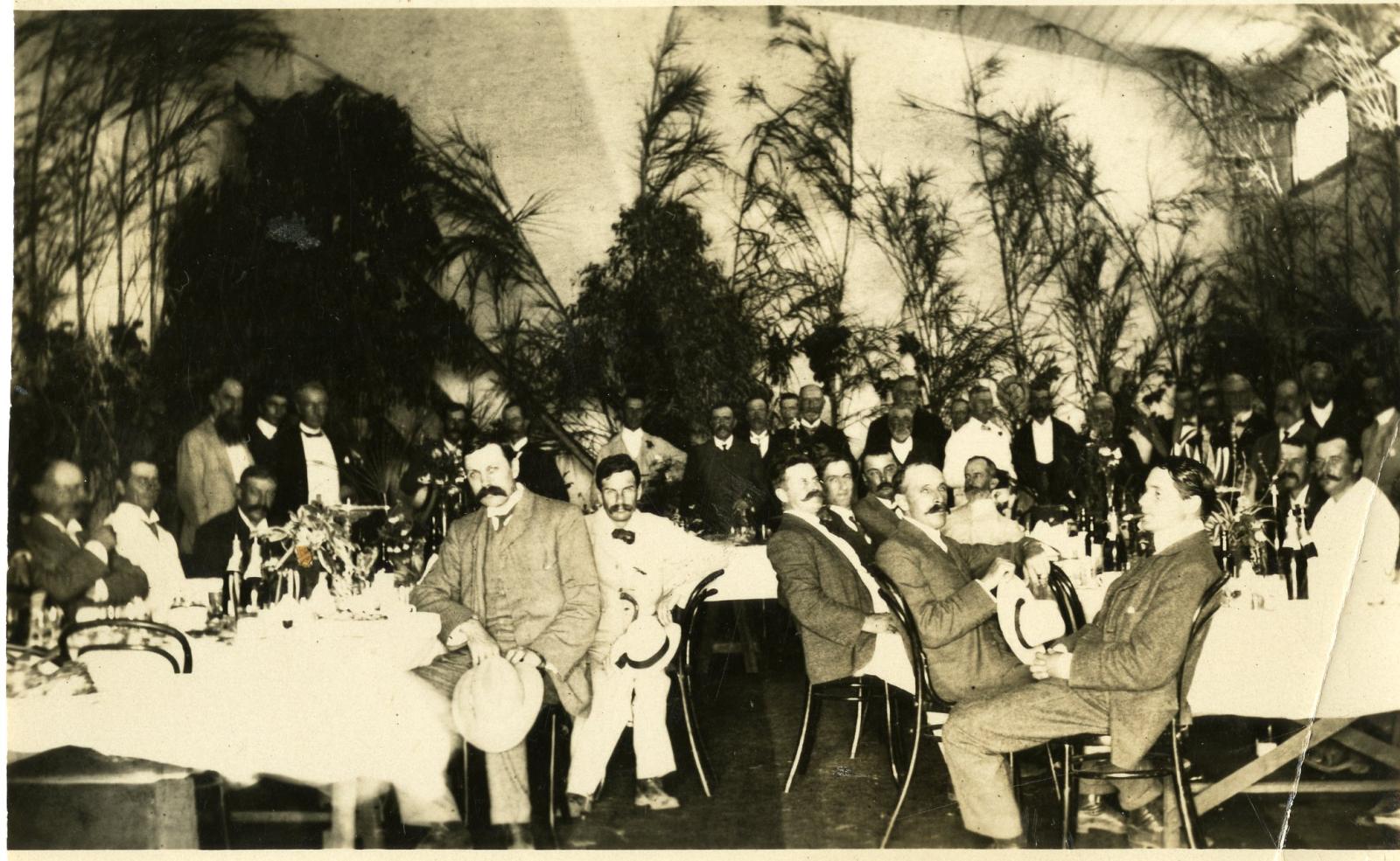 A large group of men sitting at tables, all wearing suits. The men at the back are standing so they can be seen. All are looking towards the camera