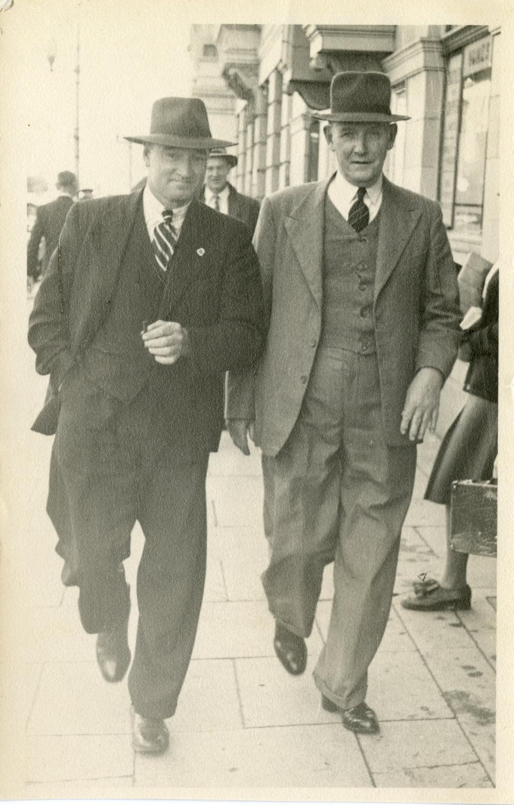 Two men walking side by side towards the camera on a city footpath, the man on the left is in a dark suit with a hat, the man on the right is wearing a lighter suit with a hat.