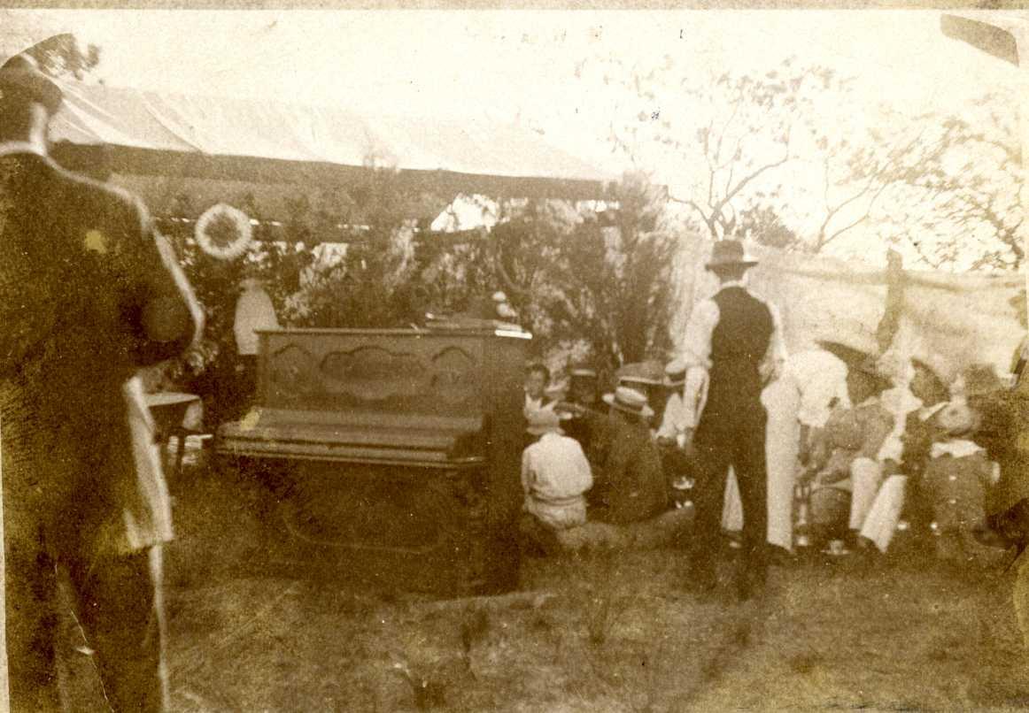 Sepia toned photograph of a upright piano sitting outside on grass with people standing around in the background. Men in pants, white shirts, vests and wearing hats, women in blouses and dresses. 