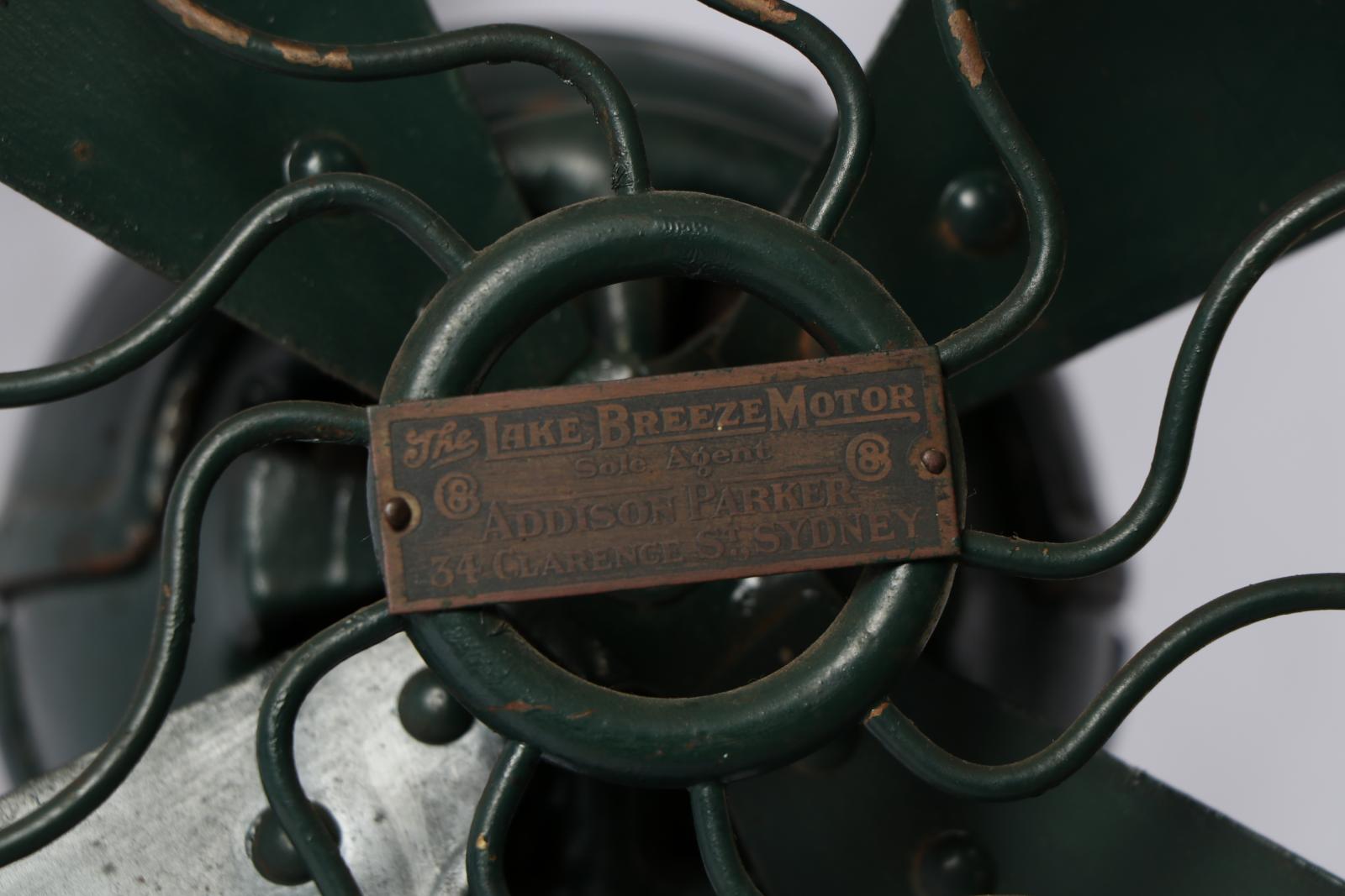 Close up of makers plate on centre front of fan, attached to green circle and squiggly green metal fans out from it.