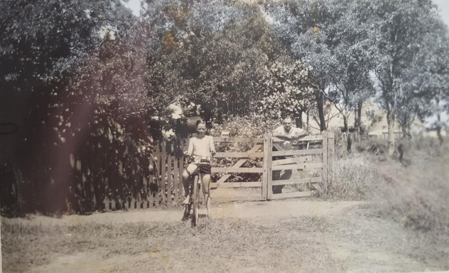 Young Dennis Walker on his pushbike, Lew Whiteman standing behind the gate, 1946/1947.