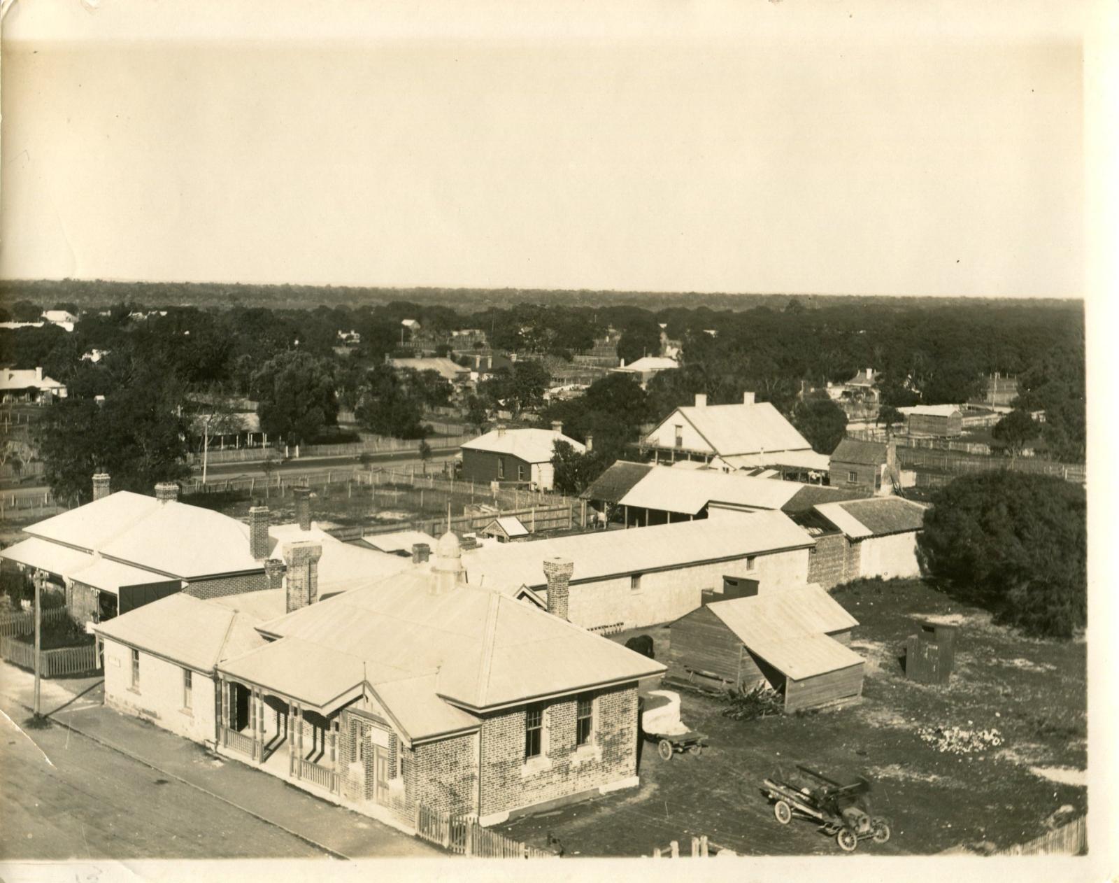 Sepia photograph of the old courthouse complex in Busselton. On the left of image is small brick building with windows, behind is a long rectangle building for the jail cells. on the left is a smaller timber building that was the Bond store. on the furthest left is a pile of grass and dirt. A car is entering from the left. 