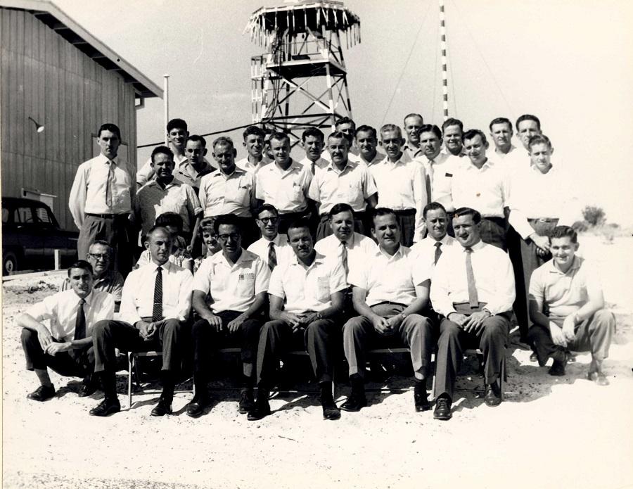 Black and white photograph of a group of men wearing short sleeve white shirts, ties and trousers with an antenna in background