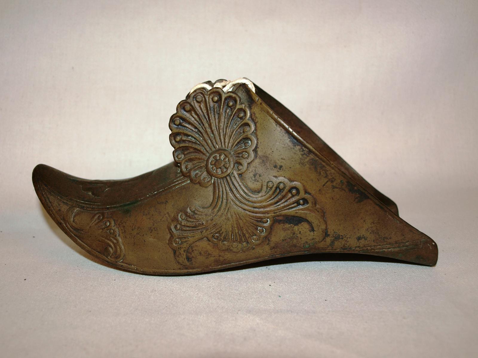 Brass, shoe-shaped stirrup on white background. Photo is showing left hand side of stirrup, which has a pointed toe and floral pattern.