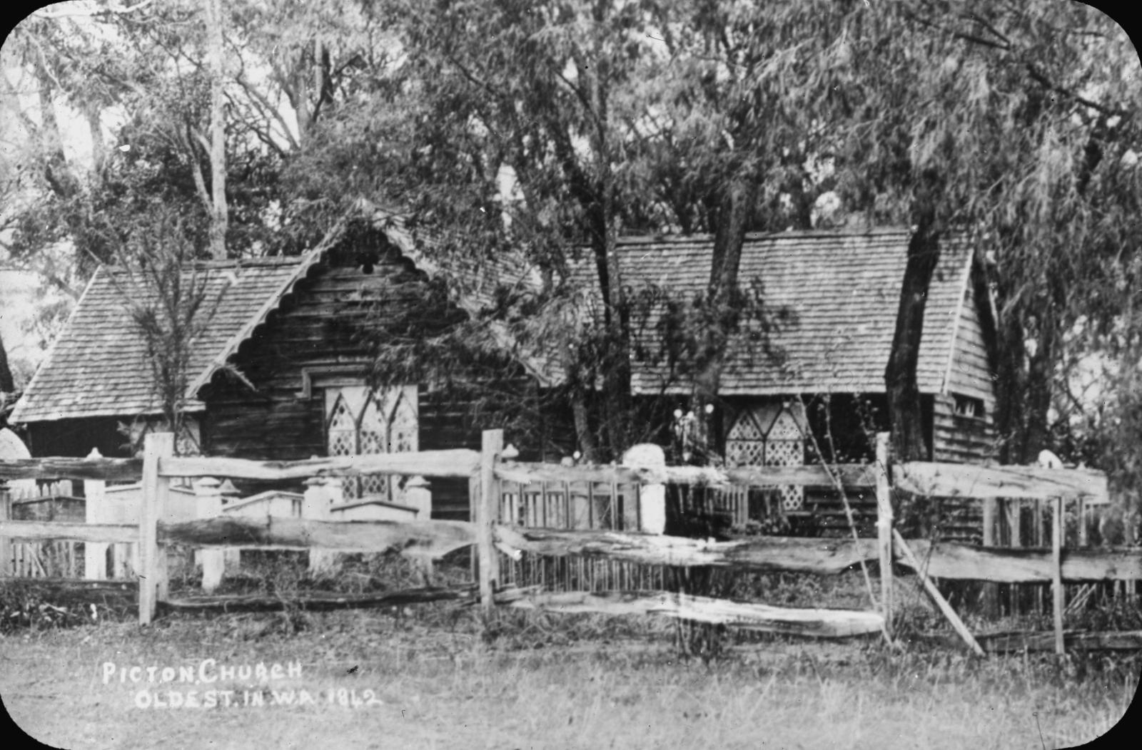 Photo of Picton Church, one of the oldest churches in the Swan River Colony.  The church is a wooden structure surrounded by a wooden fence.  Two stain glass windows are at the front of the church.  There are large trees which are close to the building.