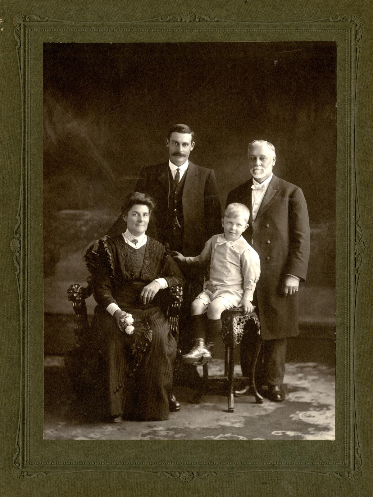 Family black and white studio portrait. Two men standing in background, wearing suits, man on left has dark hair and moustache, man on right white hair with moustache and beard. Front row is a woman, seated in a dark long sleeve, ankle length dress. To the right is a young boy, seated on a stool in shorts, collared shirt and wearing a bow tie.