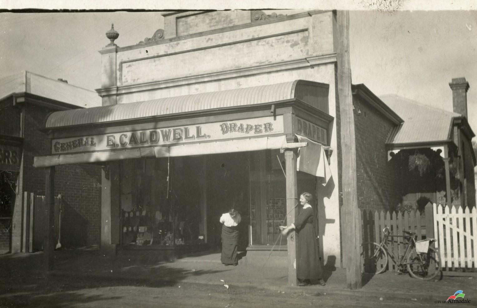 black and white photograph of a single storey shop with a verandah out front. Two women standing at the front of the shop, one looking down and wearing a dark shirt and white blouse and the other in a dark dress with arms around the right end pole of the verandah. Sign on front of verandah General E. Caldwell. Draper.