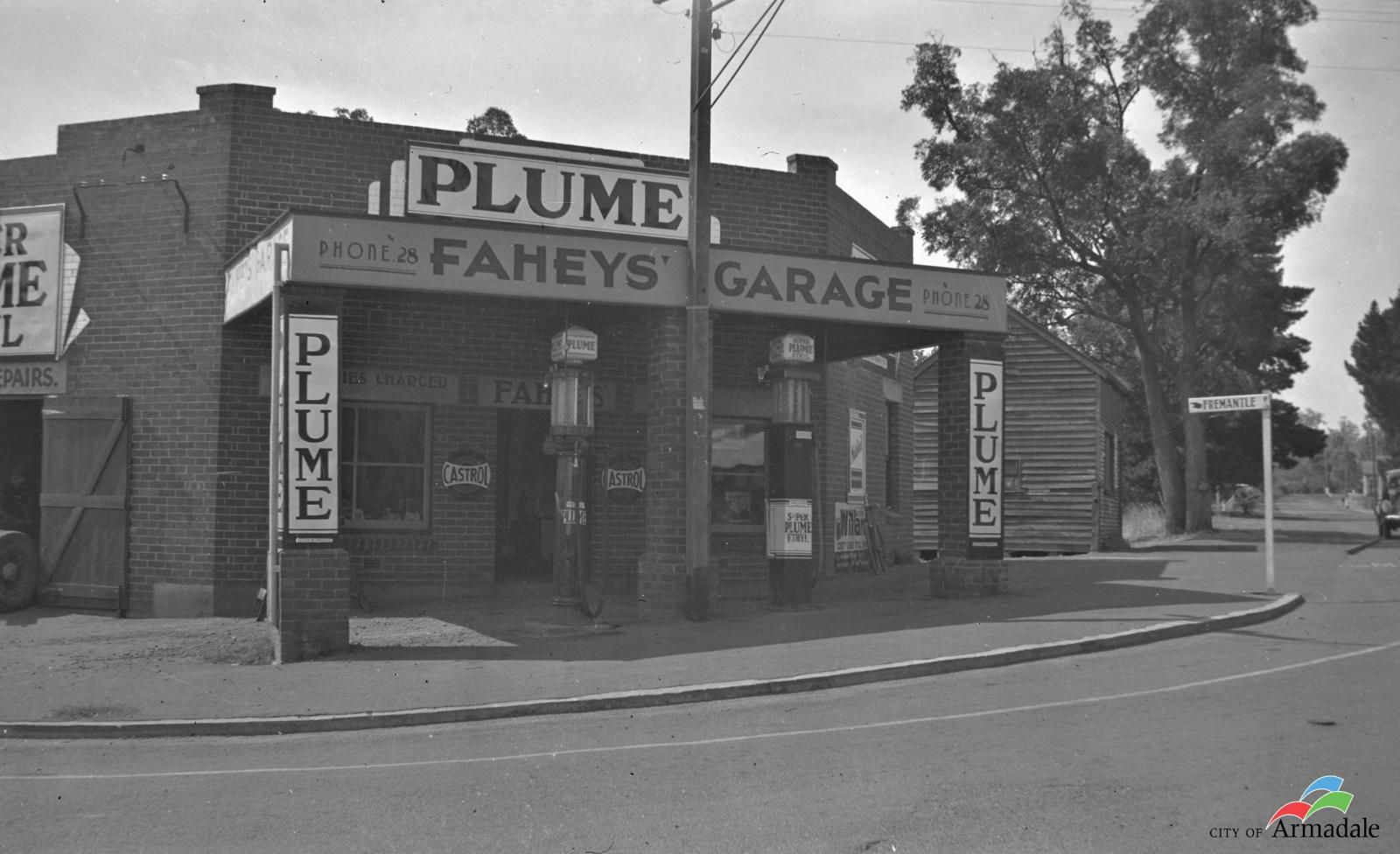 Black and white photograph of a 1930s petrol station. Front has three brick pillars and two tall petrol bowsers with glass tops and capped with cube shaped Plume signs. Metal Plume signs on end pillars and top of building. Garage name on a sign on front Faheys' Garage. Curved road in front of garage and a street sign with Fremantle and a illustration of a had pointing to the left.