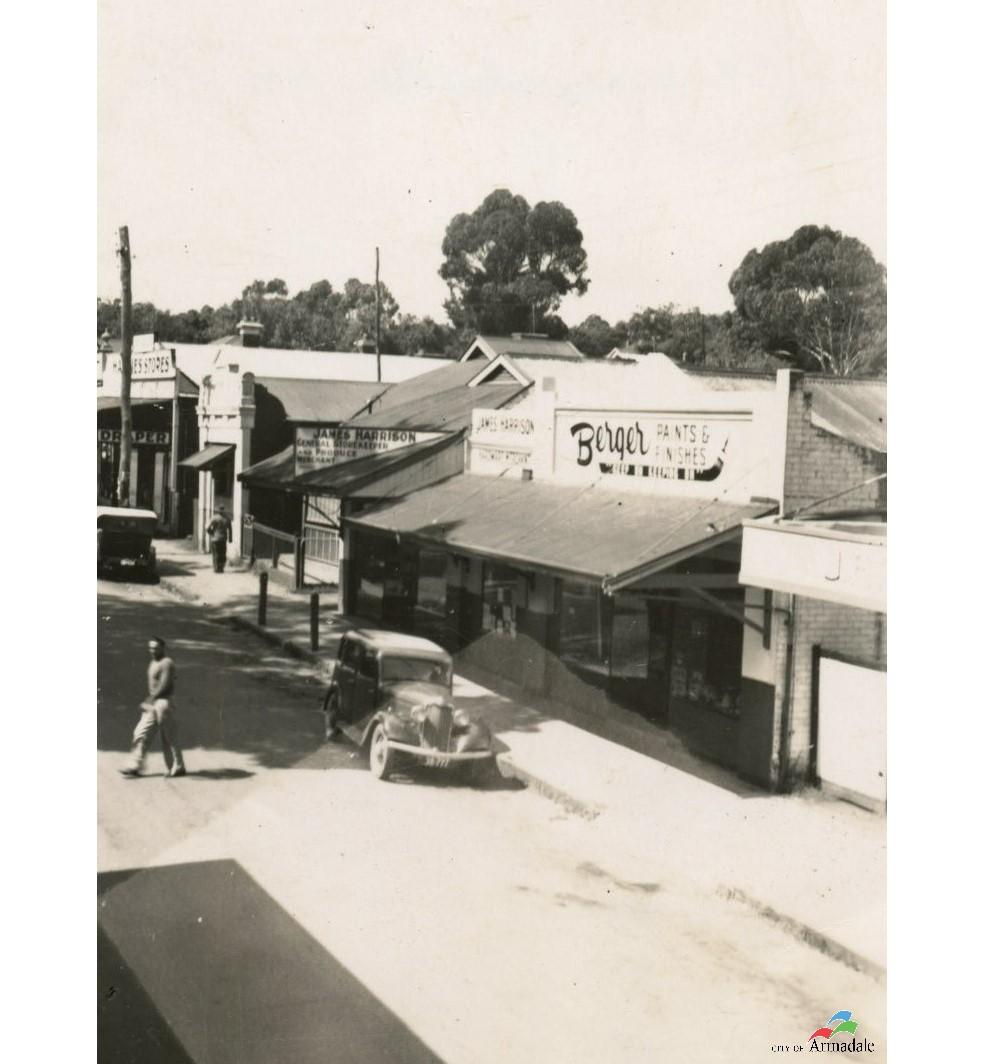 black and white photograph looking down a street. On right side are a series of shops, middle shop has a verandah at front and store sign painted above the verandah, Berger Paints & Finishes. Shop to the left has a sign saying James Harrison General Storekeeper. Car parked out the front of these shops and a man is crossing the road.