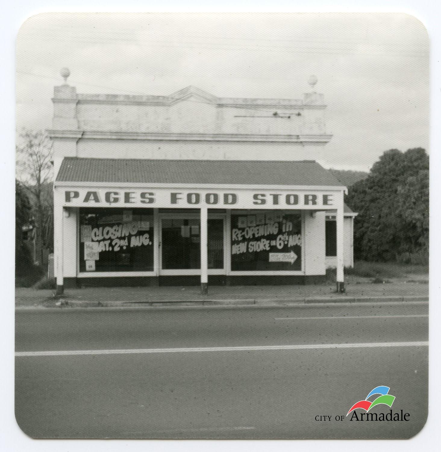 Black and white photo of front of a white single storey shop front with a veranda at front. Sign on verandah Pages Food Store. Two glass windows at front with glass door between them. Advertising text written on front. 