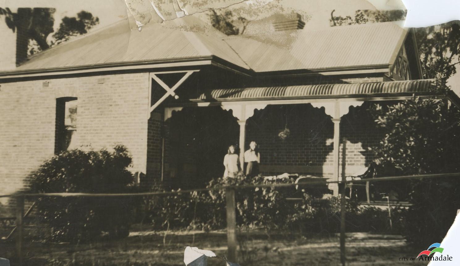 Black and white photo of front of brick house with corrugated iron pitched roof, veranda at front, two women standing on front veranda. garden with low shrubs and a fence in front of house 