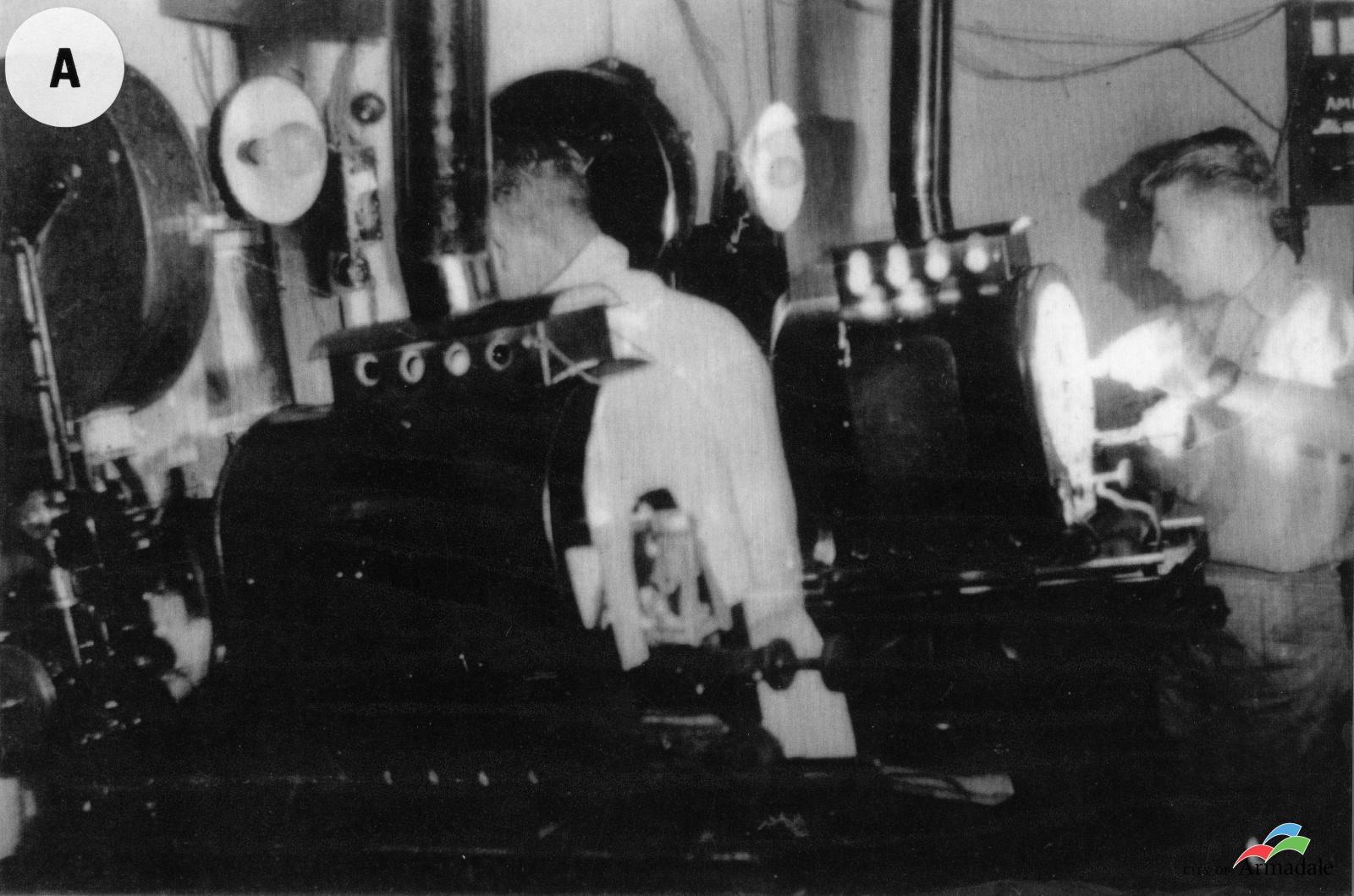 Black and White photograph of two men in a small room operating large film projectors