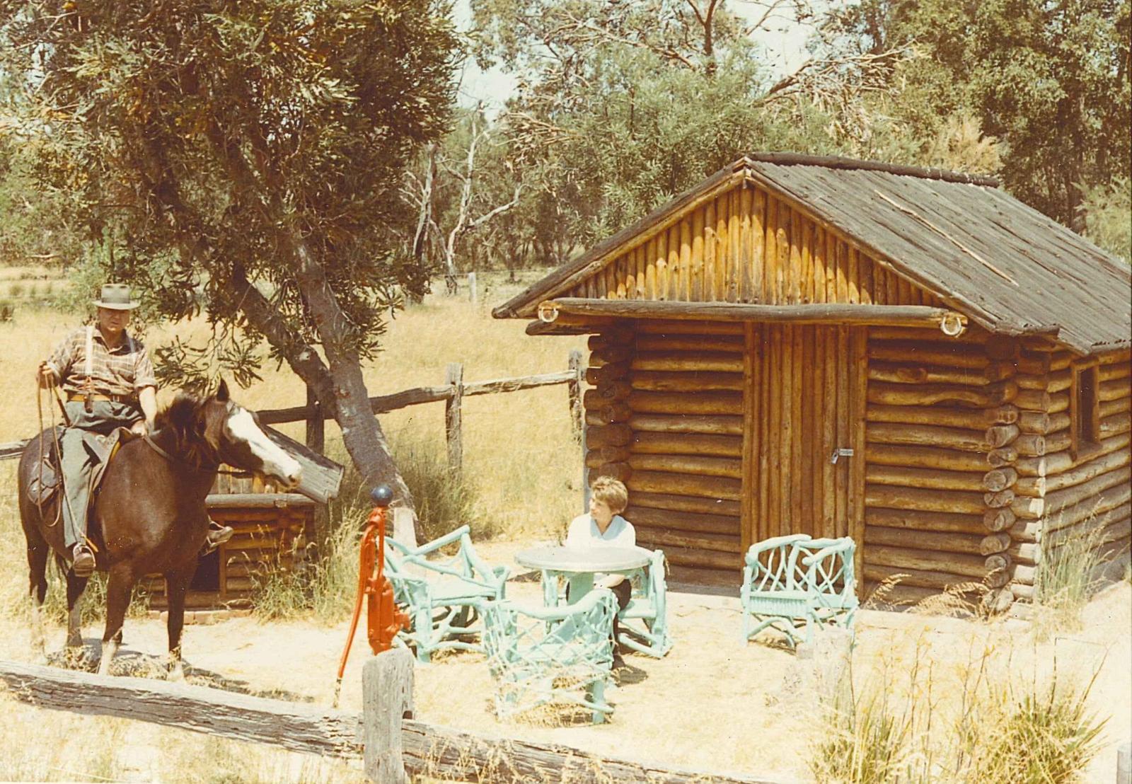 Photograph of man on horseback and woman sitting at a small blue table in front of square log cabin. Area is fenced off, with tall yellow grass and bushland in the background.