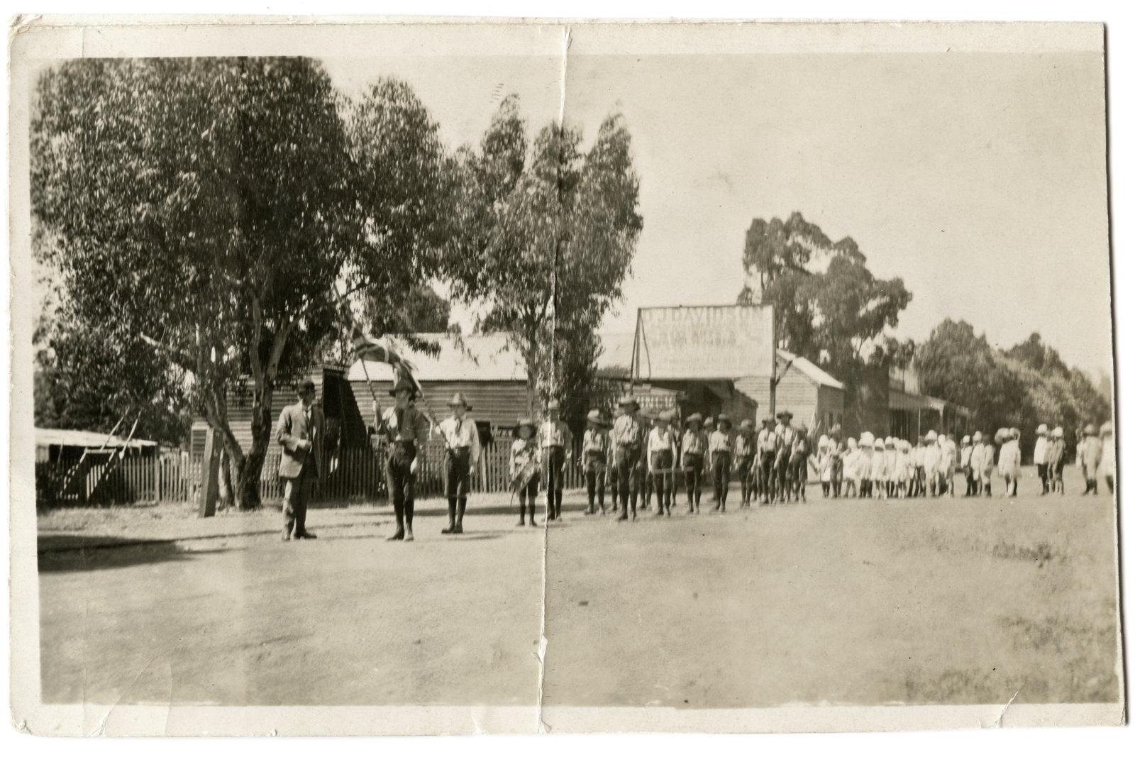 Black & white photo of boy scouts lined up on a road outside a wooden building with a tin roof. Sign outside of building says D. J. Davidson across the top