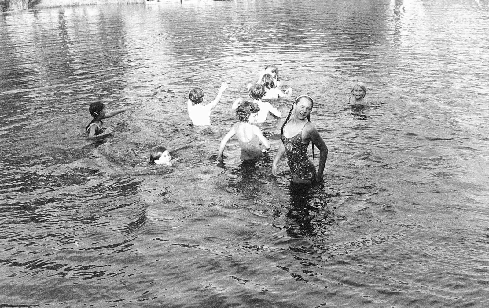 Black and white photograph of children swimming in expanse of water, which takes up entire background and foreground. A young girl stands and faces the camera, indicating that the water is shallow. 