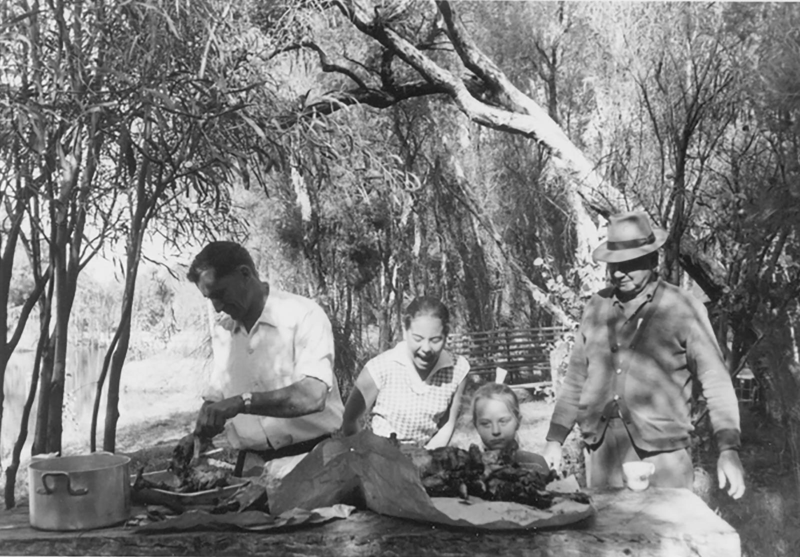 Black and white photograph of a man in a white shirt, a woman in a white shirt, a young girl and a man in a brimmed hat and cardigan setting up cooked meat on a wooden table. They are situated amongst tall trees, with a bridge in the background.