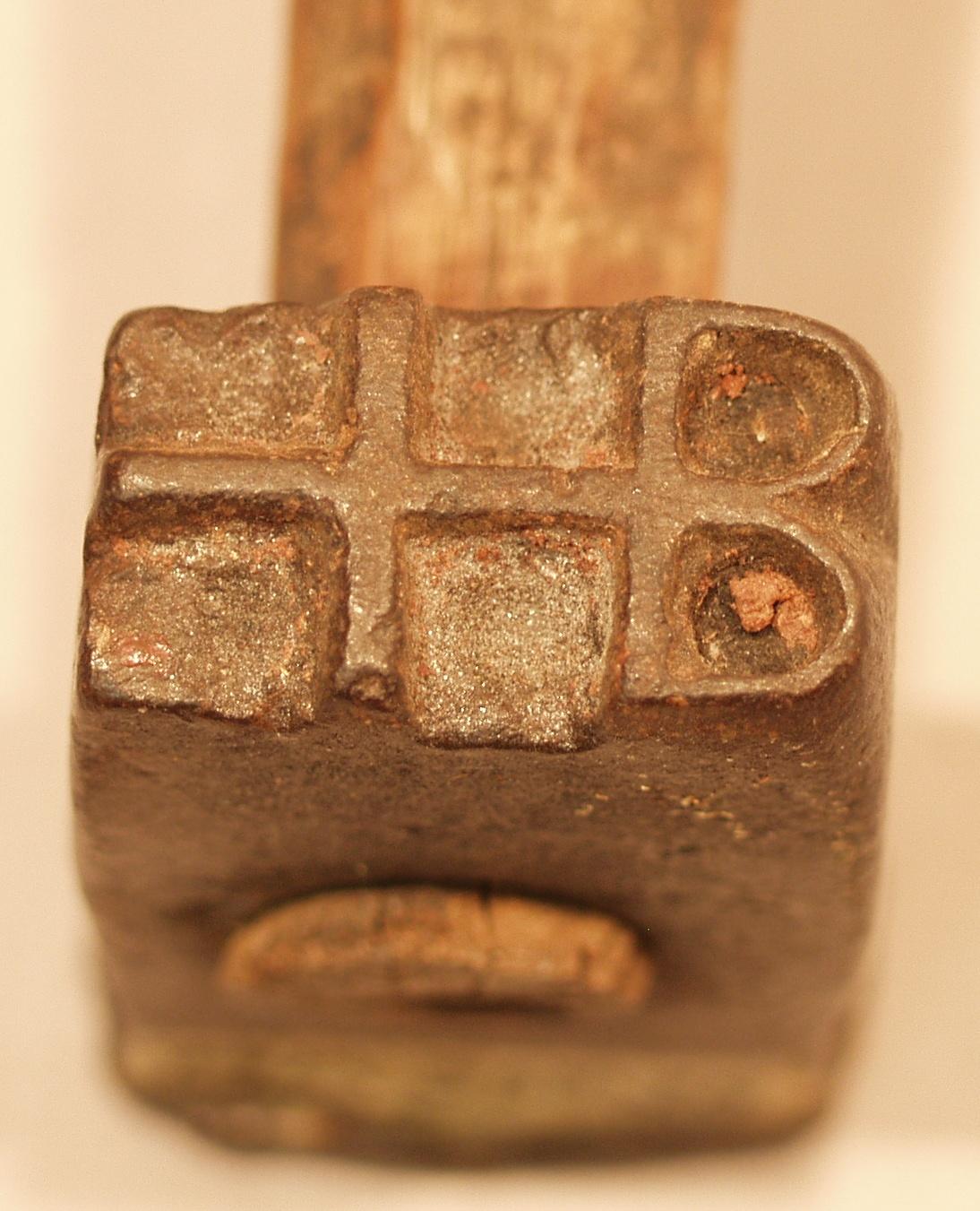 Close up view of rectangular metal head of a branding hammer showing a moulded "+B" in the metal