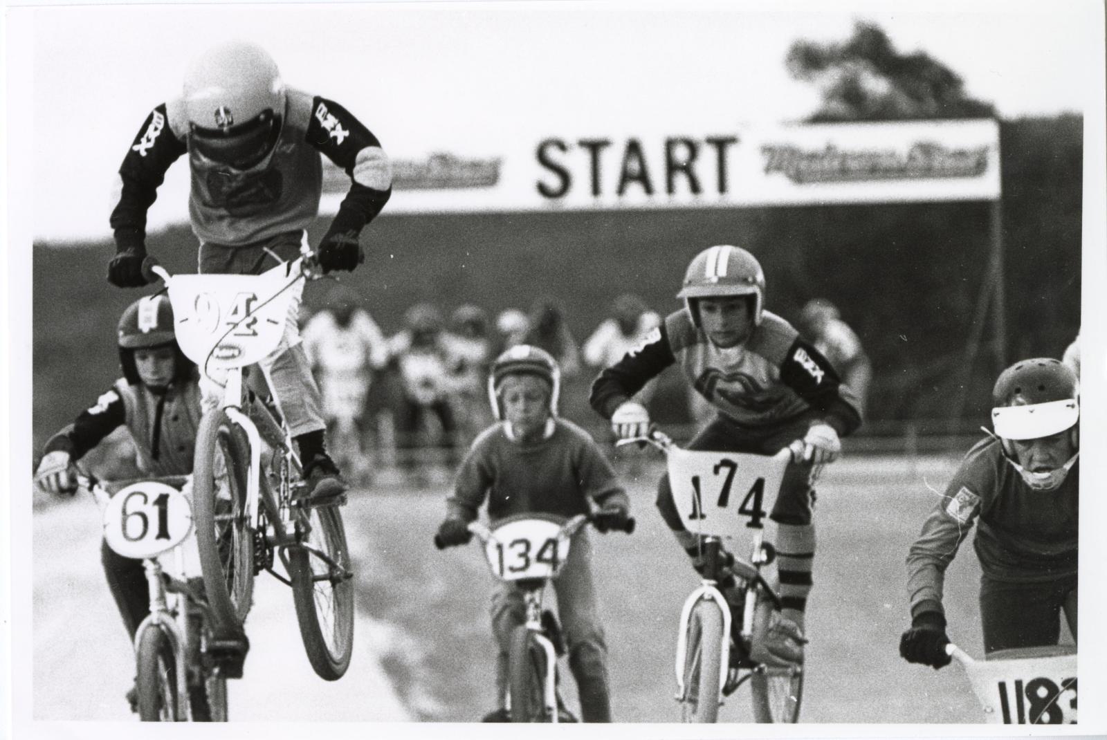 Black and White photograph of 5 teenagers on BMX bikes going over a jump. all wearing helmets and there are race numbers on the front of their bikes. In the back ground is a banner saying START