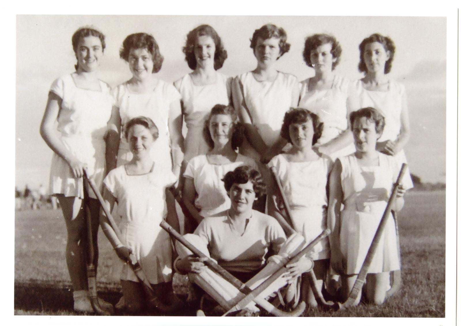 Black and white photograph of three rows of women in white uniforms. Front two rows holding hockey sticks