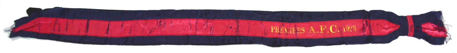 long rectnglular silk hat band made up of three horizontal stripes, dark blue, red and dark blue. At the right end in gold text 'Premiers A.F.C. 1923'