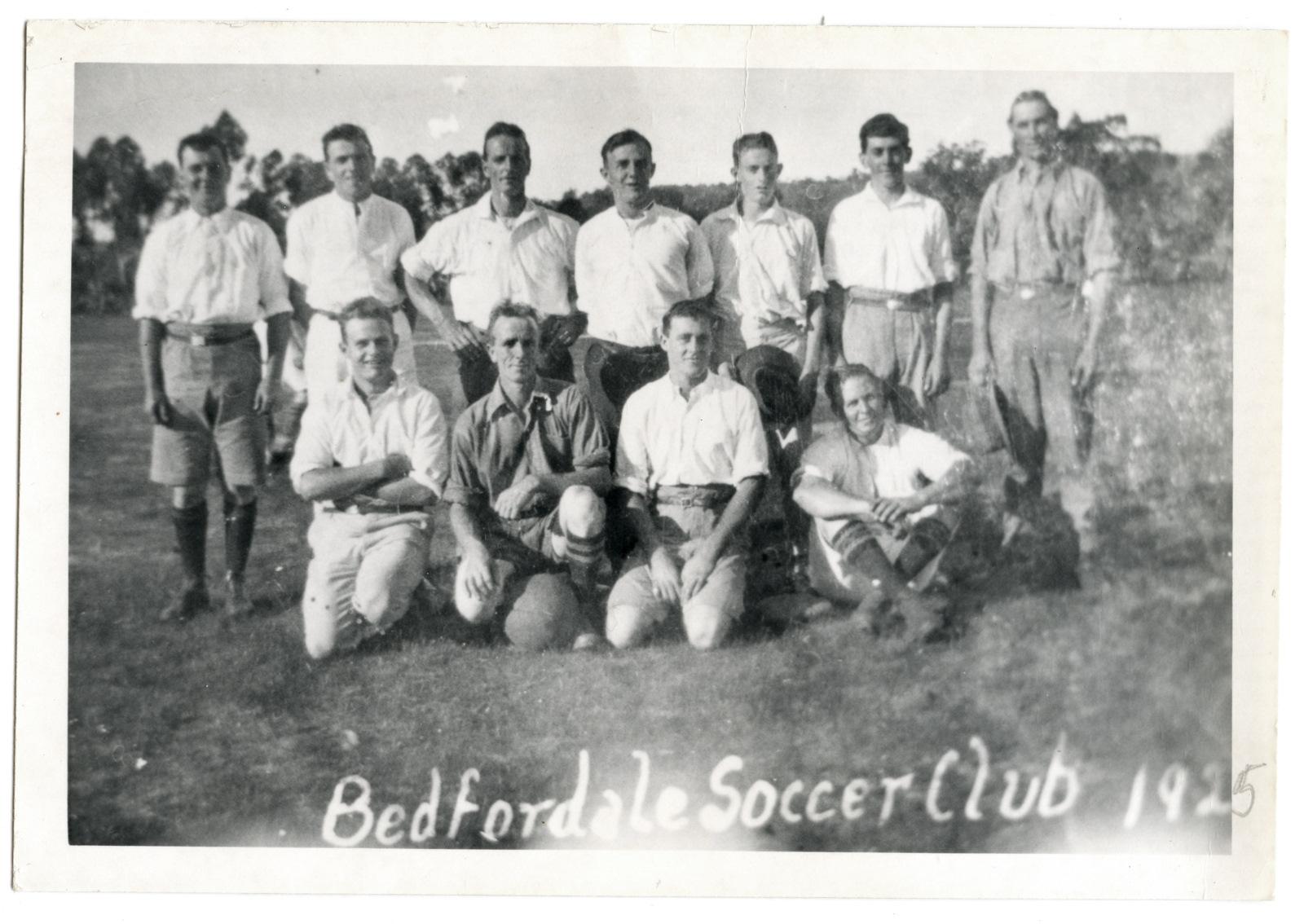 Black and white photograph of two rows of men wearing white shirts and shorts. Back row is standing, front row is kneeling. One man in the front has a round football / soccer ball in front of him. Text written across bottom of photograph 'Bedfordale Soccer Club 1925'
