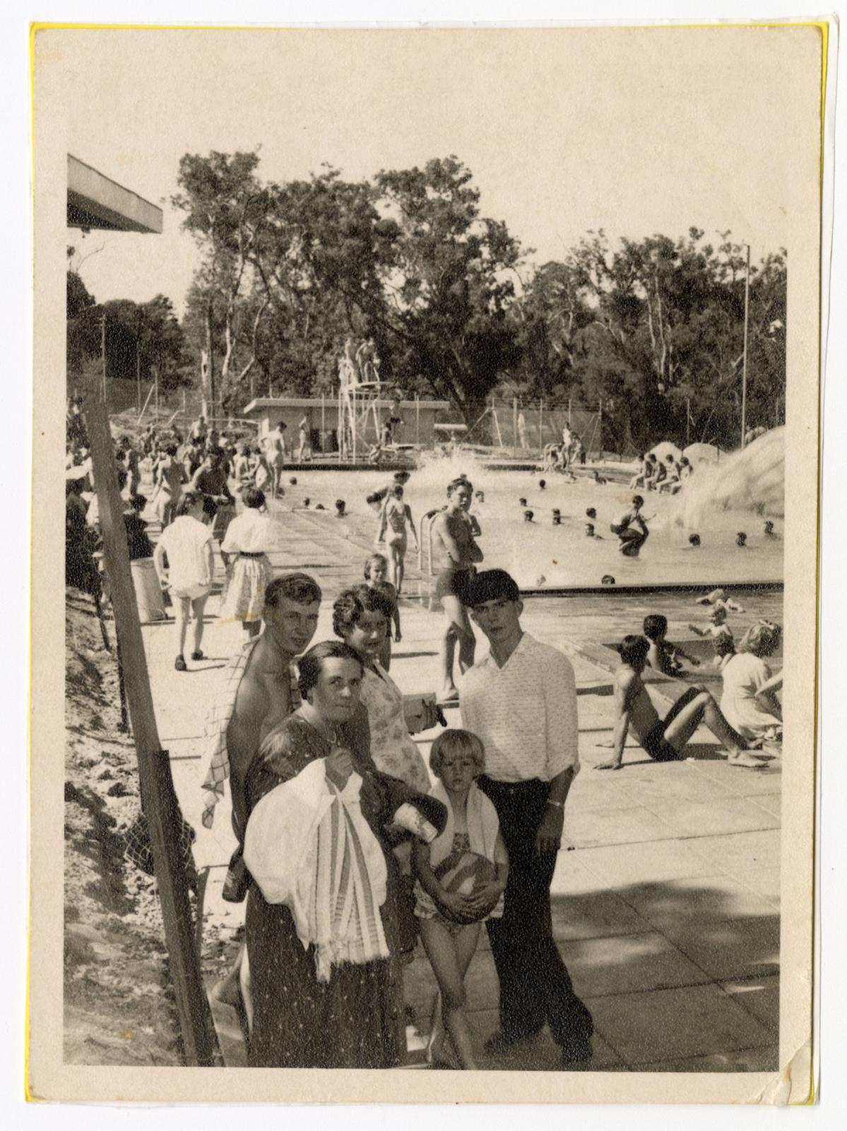 black and white photograph of a family posing in front of a public pool. In background people are swimming in the pool. A diving board can be seen at the far end of the pool.