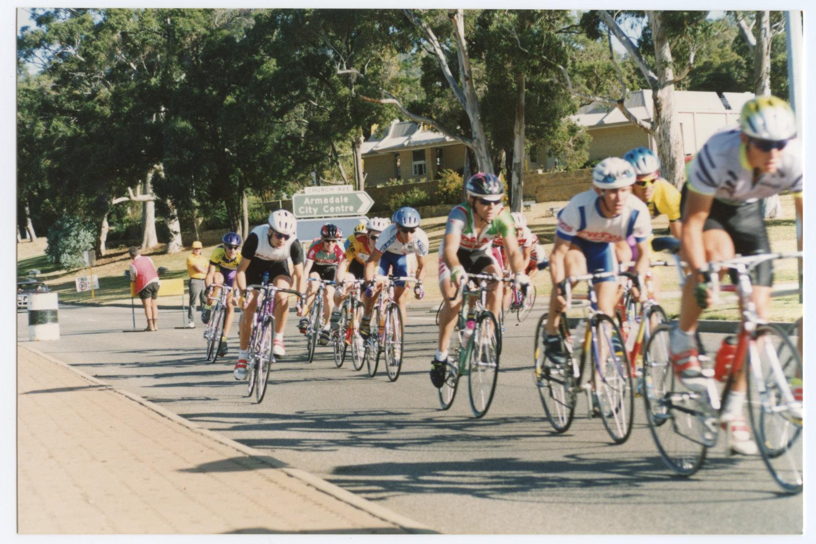 Colour photograph of cylisits on road racing bikes and wearing cycling racing uniforms riding on a paved road, trees and buildings in background