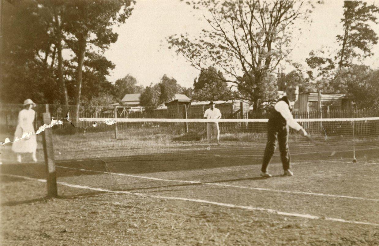 black and white photograph of two women and one man standing on a grass tennis counrt in 1925. The man is searving the ball and is wearing dark trousers, white shirt and a dark waistcoat