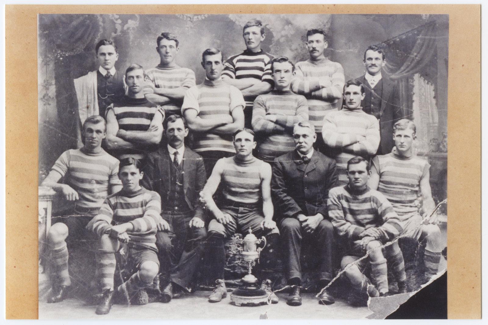 Studio black and white photograph members of the Kelmscott Football Club. The men are in three rows, two standing and front row sitting. Most are wearing shorts and horizontally striped jerseys. In front of the first row is a metal cup or trophy.