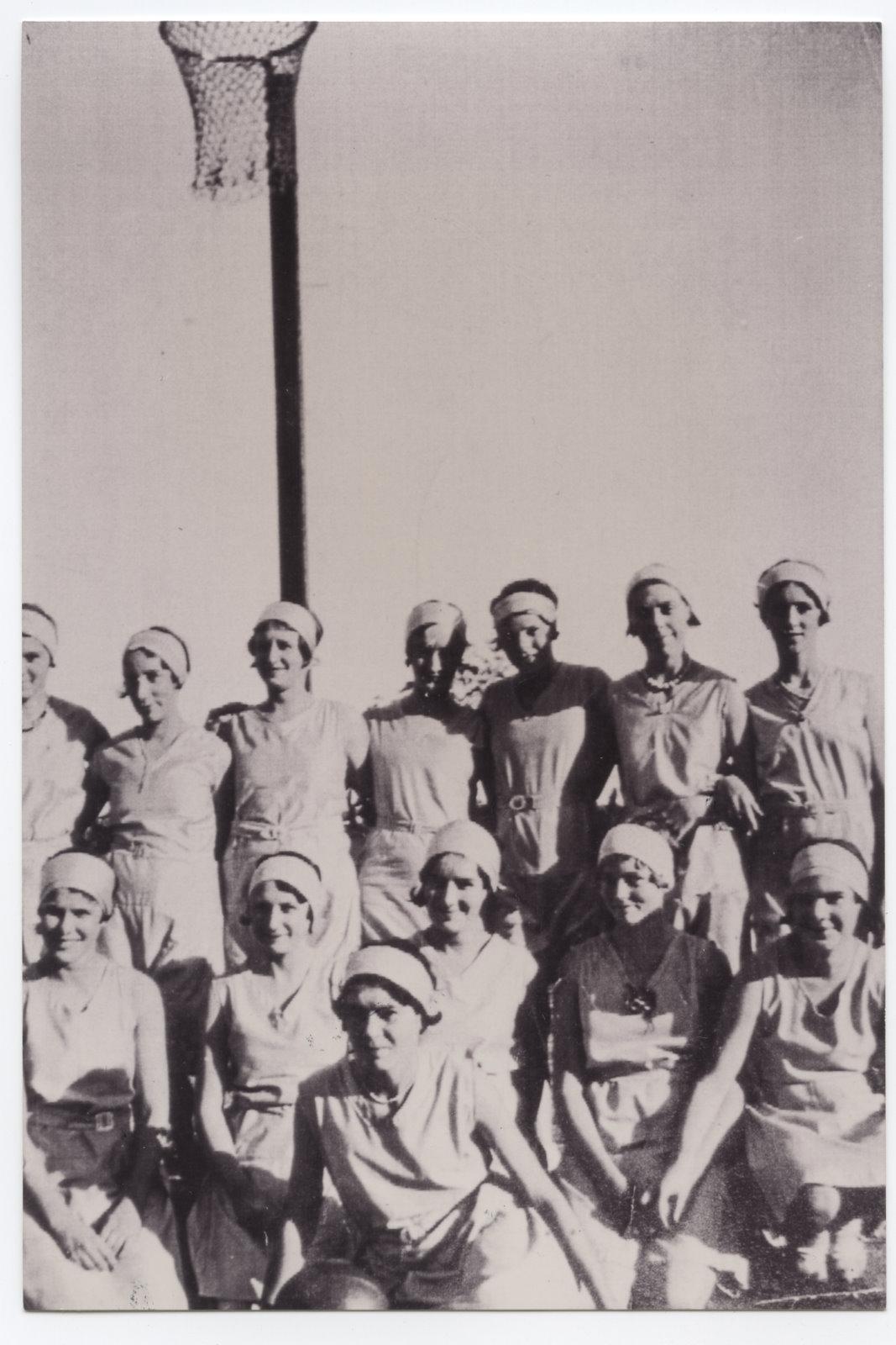 black and white photograph of a group of women posing. All are wearing a light coloured sports uniform and are wearing a head band. Behind them is a netball hoop on a pole.