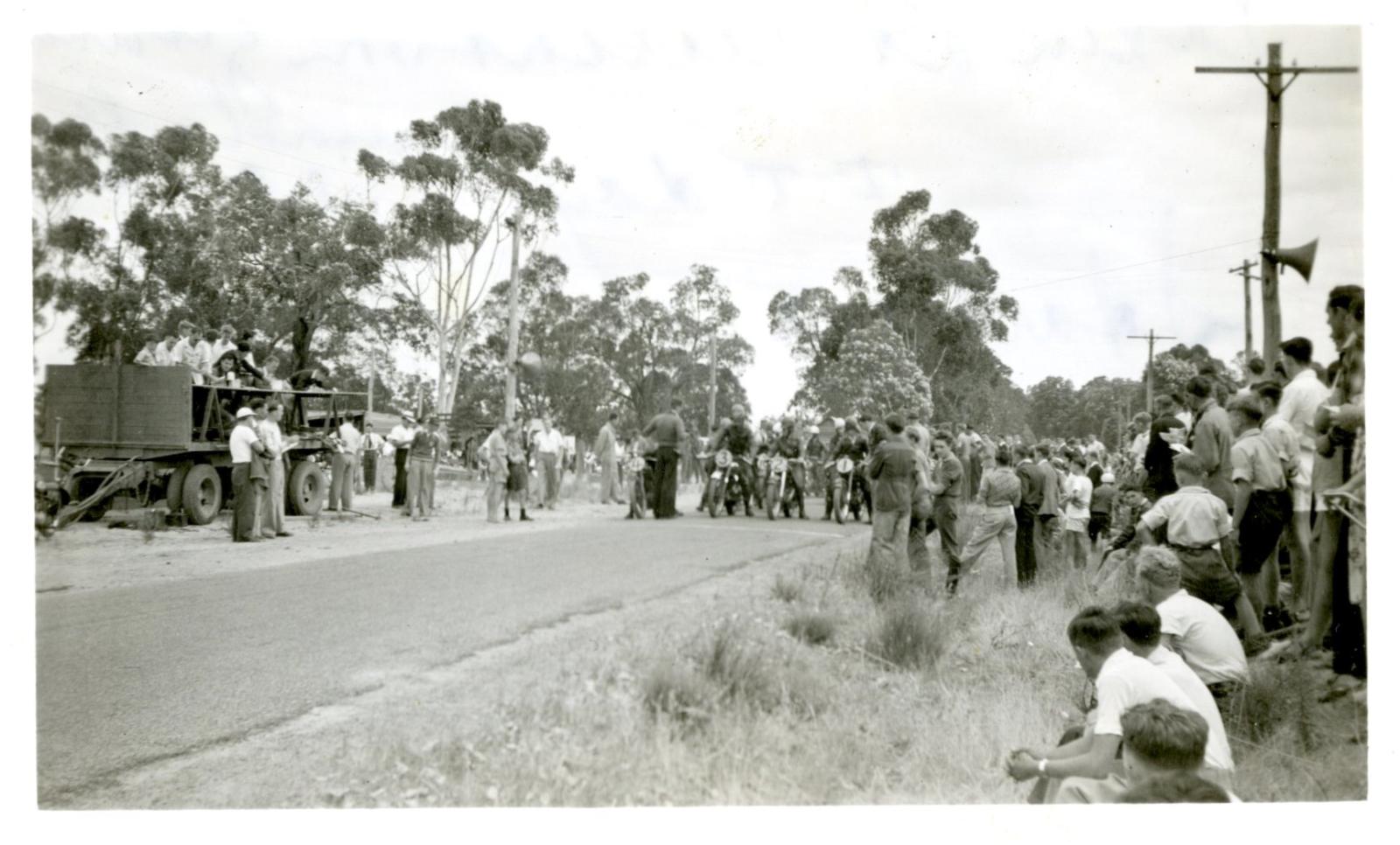 black and white photograph of a paved street with people down both sides. At the end of the street are a row of figures on motorbikes getting ready to race