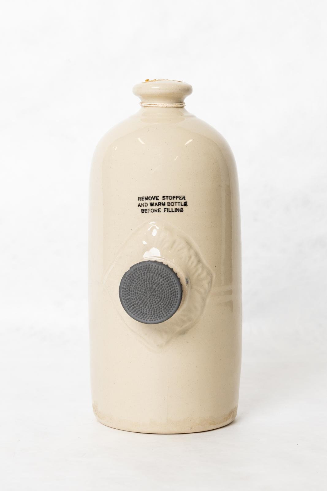 Stone coloured, ceramic hot water bottle. Rounded with a flat base and tapered narrow neck. Rubber stopper in the centre of the bottle with a moulded diamond pattern around the stopper.