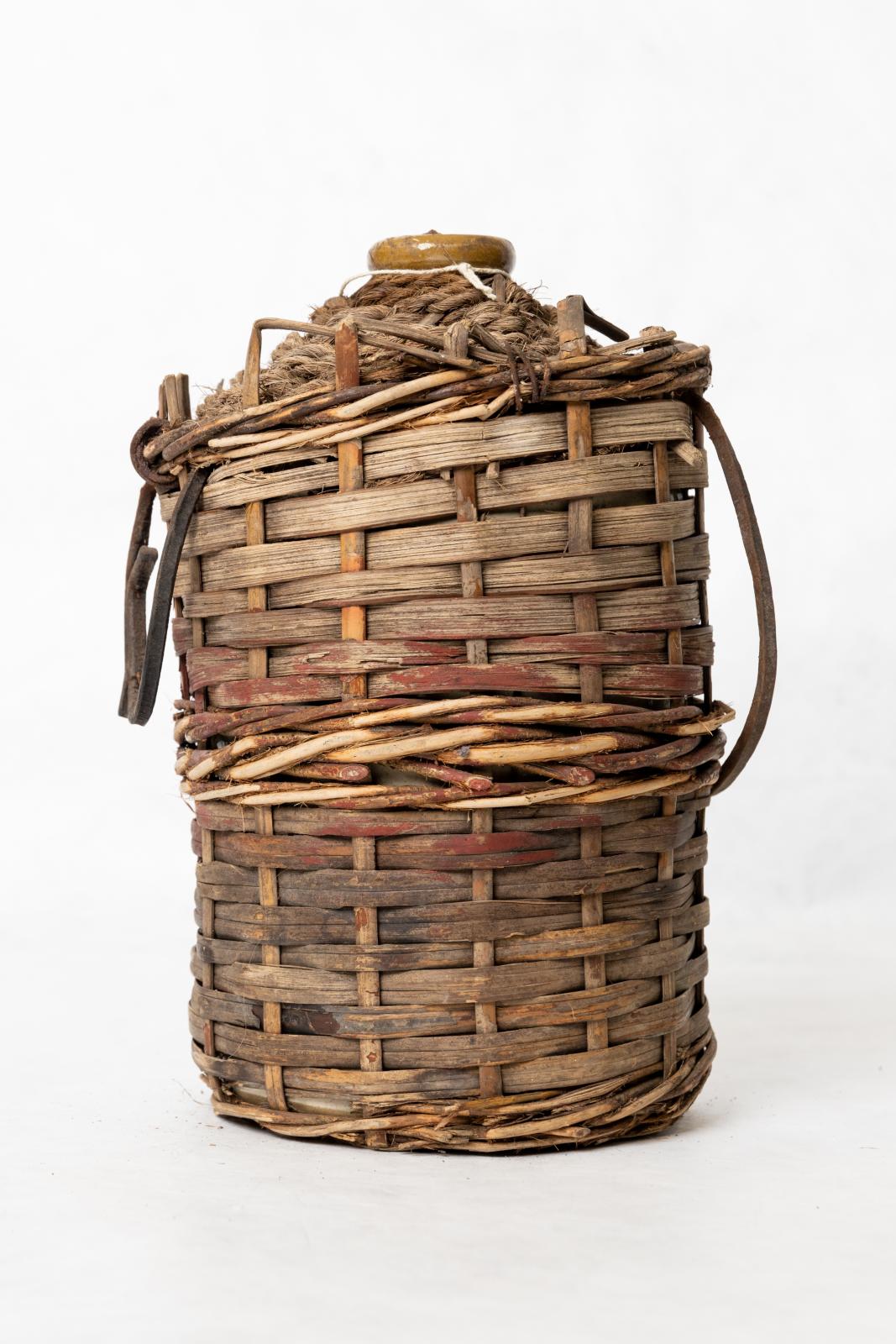 Colour faded, brown and red painter wicker jug cover. There is rope wrapped around the neck of the bottle, only the ceramic spout is visible.