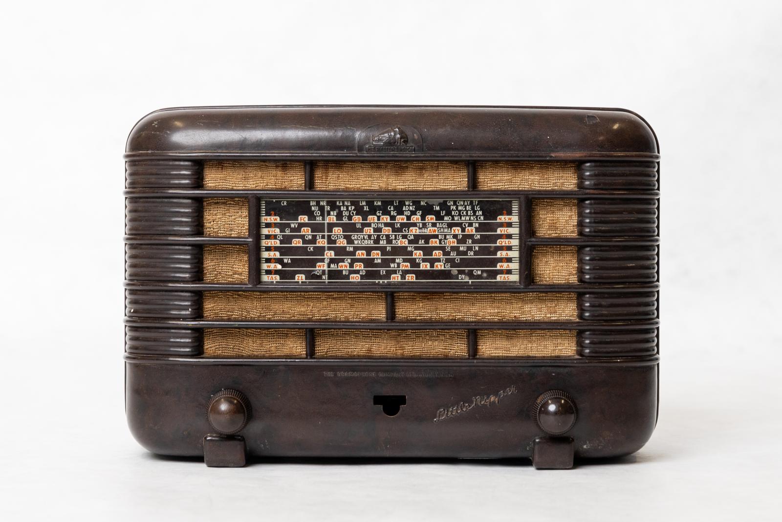 Dark brown, rectangular shaped, short wave mantle radio with station selector in the centre on top of open rectangular grid filled with light brown cloth.  Two small brown knobs, one located on the left and the other on the right. 