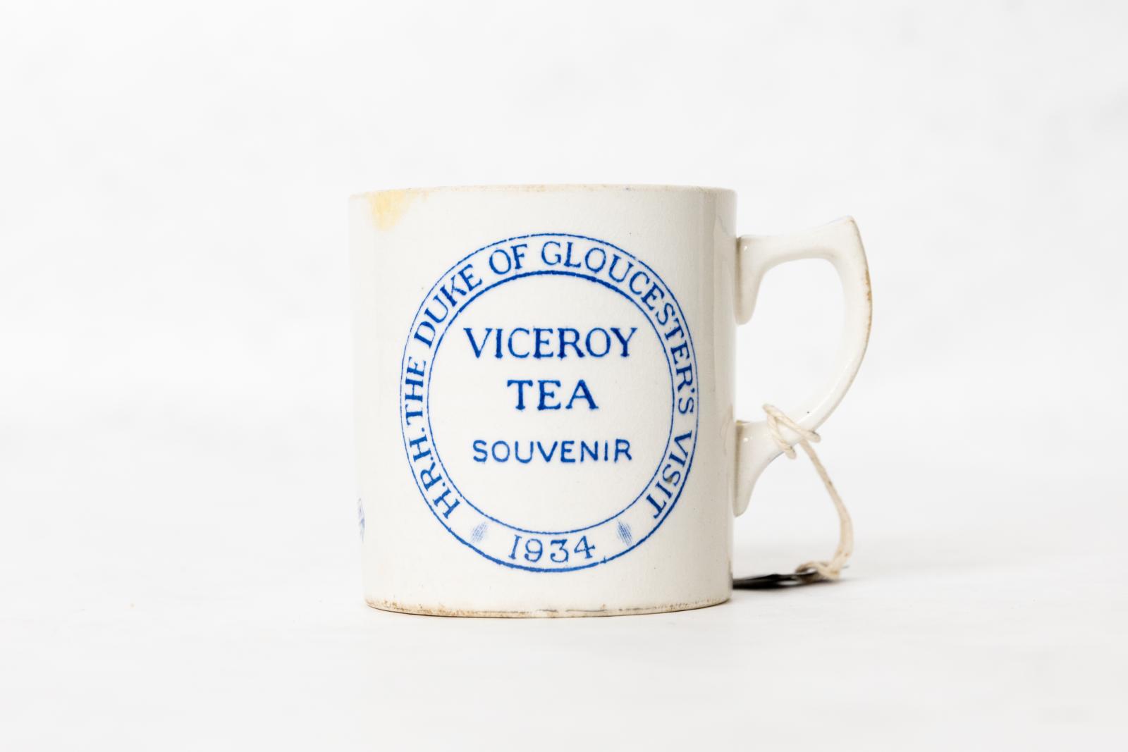 White ceramic souvenir mug with crazed glaze and blue transfer text "H.R.H THE DUKE OF GLOUCESTER'S VISIT' '1934' within circular bordered lines. In the centre of the circle is the text 'VICEROY TEA SOUVENIR'.