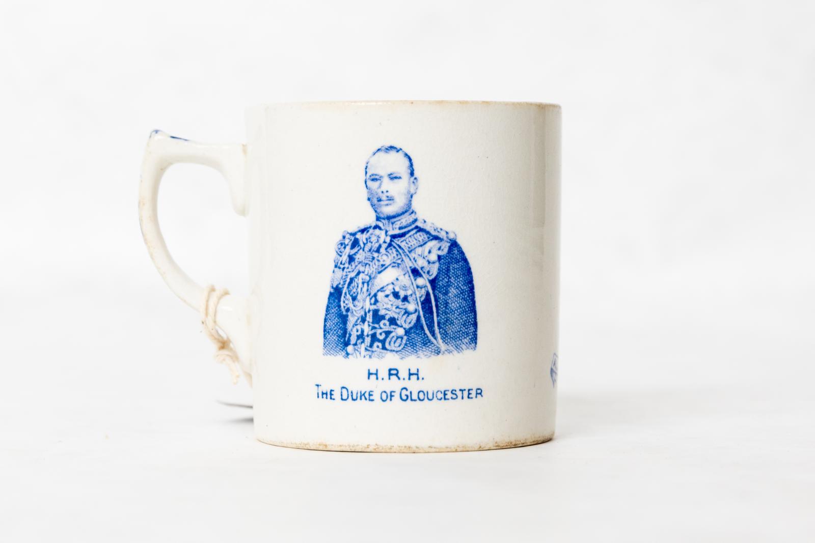 White ceramic souvenir mug with crazed glaze and blue transfer, photographic impression torso portrait of the Duke of Gloucester in his military regalia, with the text 'H.R.H. THE DUKE OF GLOUCESTER' underneath the image.