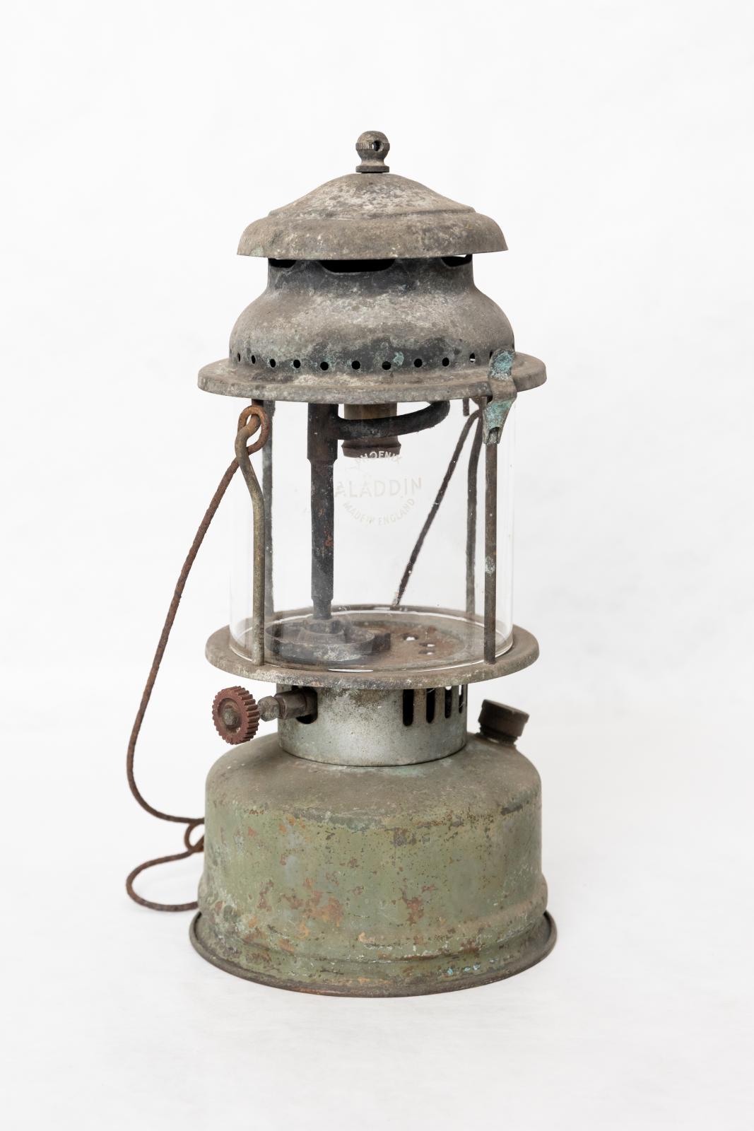 Large metal pump kerosene/ petrol lamp. Brass plated and painted green, the paint is peeling. The base/ kerosene tank has a brass pump, brass screw cap and knob to adjust the flame. Metal stands to support gallery and encase glass, the gallery is eroded. There is a metal swing handle with hook for suspending. 