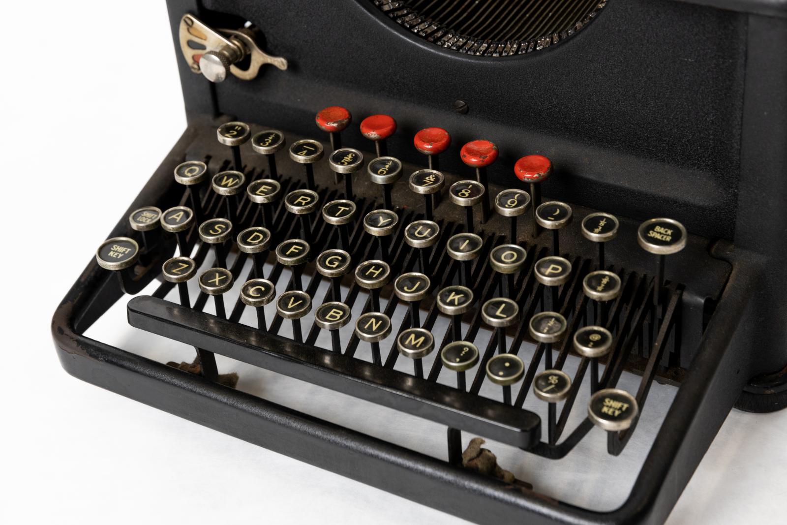 Textured black metal "REMINGTON" office typewriter with four banks of circular keys with white letters on black background framed with chrome. Chrome carriage return lever and tab measures. Geared type bar mechanism with simple linkage between type lever and type bar. "Remington" gold lettering on paper support with image of typewriter in centre of red circle.