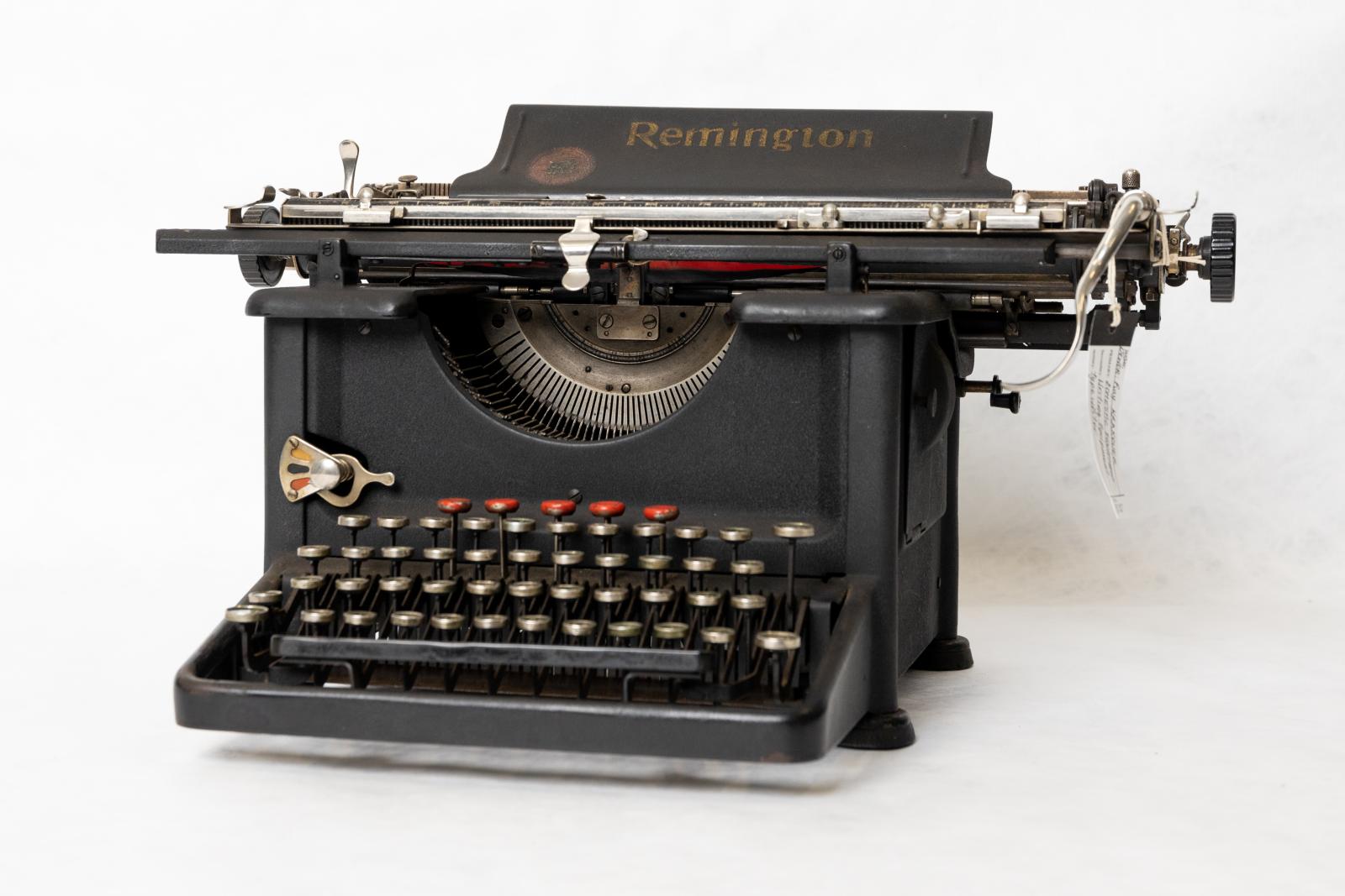 Textured black metal "REMINGTON" office typewriter with four banks of circular keys with white letters on black background framed with chrome. Chrome carriage return lever and tab measures. Geared type bar mechanism with simple linkage between type lever and type bar. "Remington" gold lettering on paper support with image of typewriter in centre of red circle.