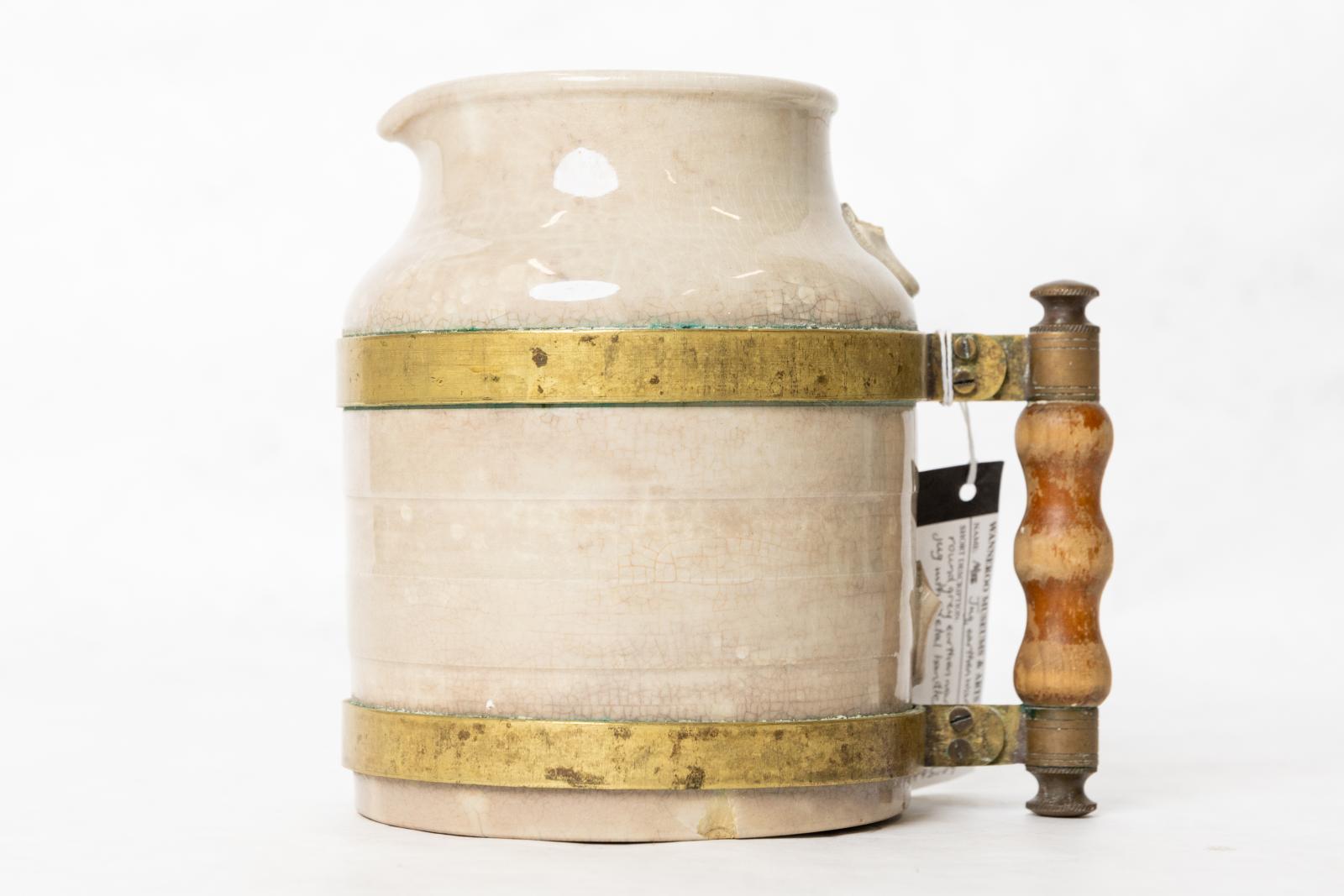 Round grey earthenware milk jug, with the original handle broken off and a metal and wooden handle attached with two brass rings around middle of jug.