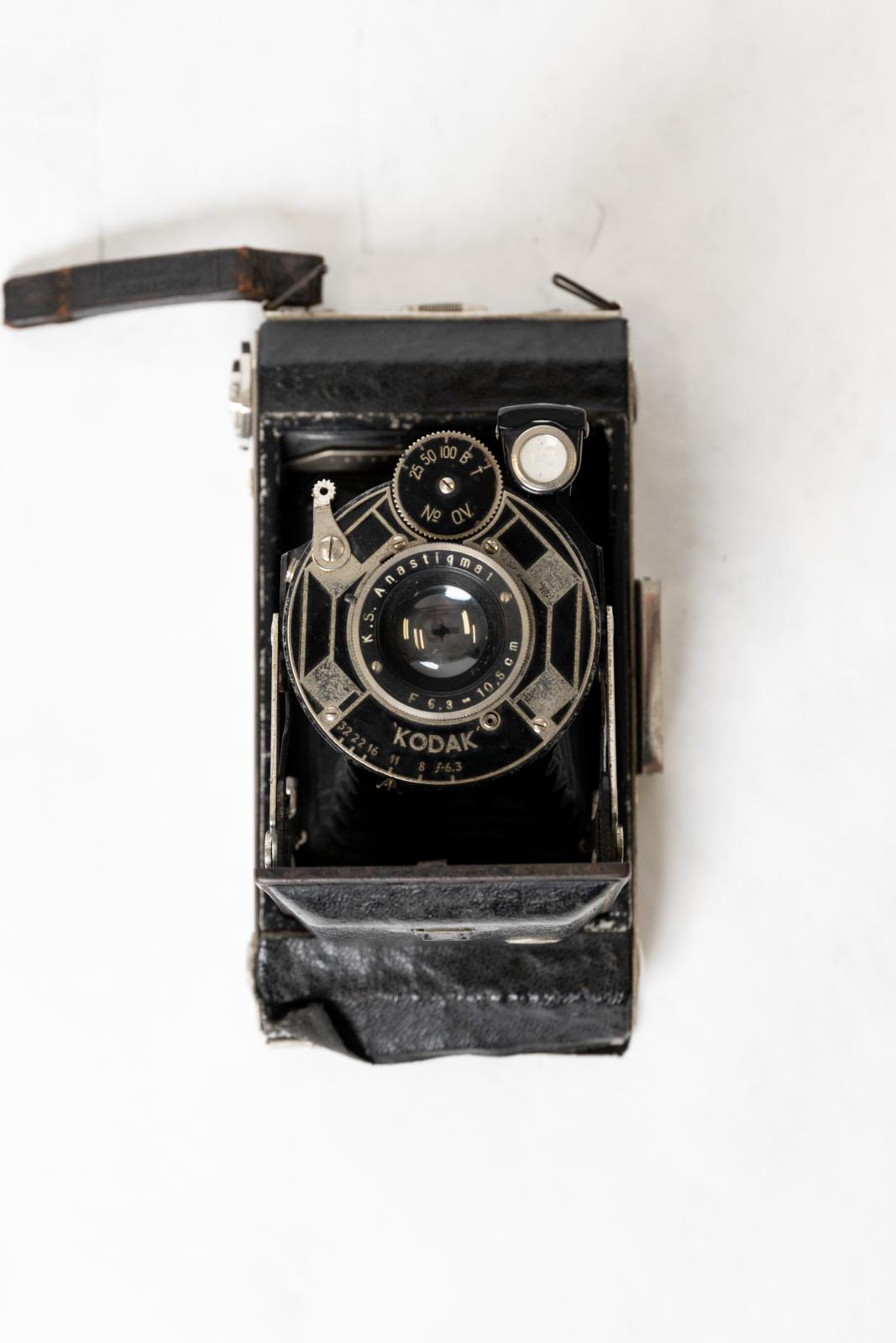 Rectangular, black metal and cloth, Kodak camera. The lens folds out on a concertinaed cloth sleeve held by two metal arms resting on a square metal plate. There is a small square viewfinder on the top and on the front a round glass viewfinder and small round black dial with numbers, a small silver metal shutter release and a round silver metal lens. The entire lens folds in neatly and covered by the metal plate. On the base there is a metal winder and on the top a square metal box with a viewfinder.