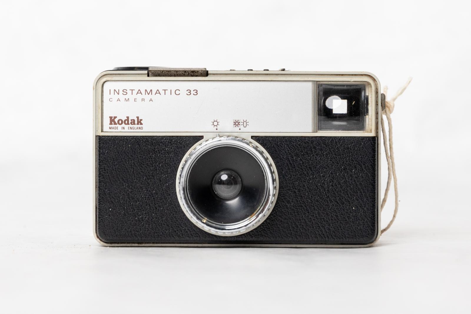 Rectangular metal box with black leather covering. On the front is a round metal and glass lens, with a glass viewfinder in the top right hand corner. It is surrounded by plastic and the front panel is aluminium.