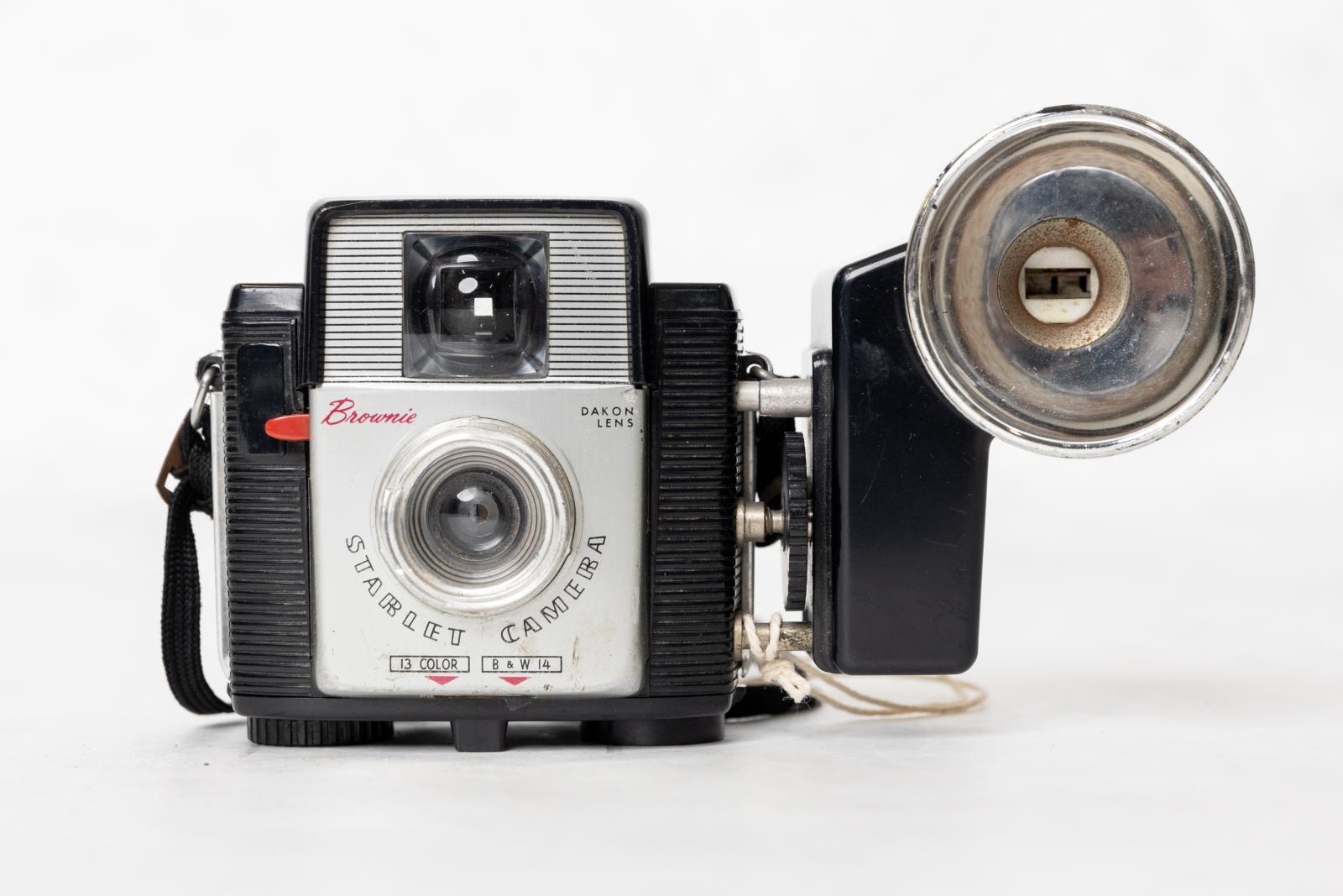 Rectangular black plastic box with a silver metal panel on the front with a round metal lens, a square glass viewfinder and a red plastic shutter release knob. On the base there are two round plastic knobs, one is the winder and the other the lock. On the top there is a silver metal plate and at the back there is a small square glass viewfinder with a small round red plastic knob. On each side there is a small fitting which holds a cloth shoulder strap. Mounted on the right side is a flash, black plastic wa